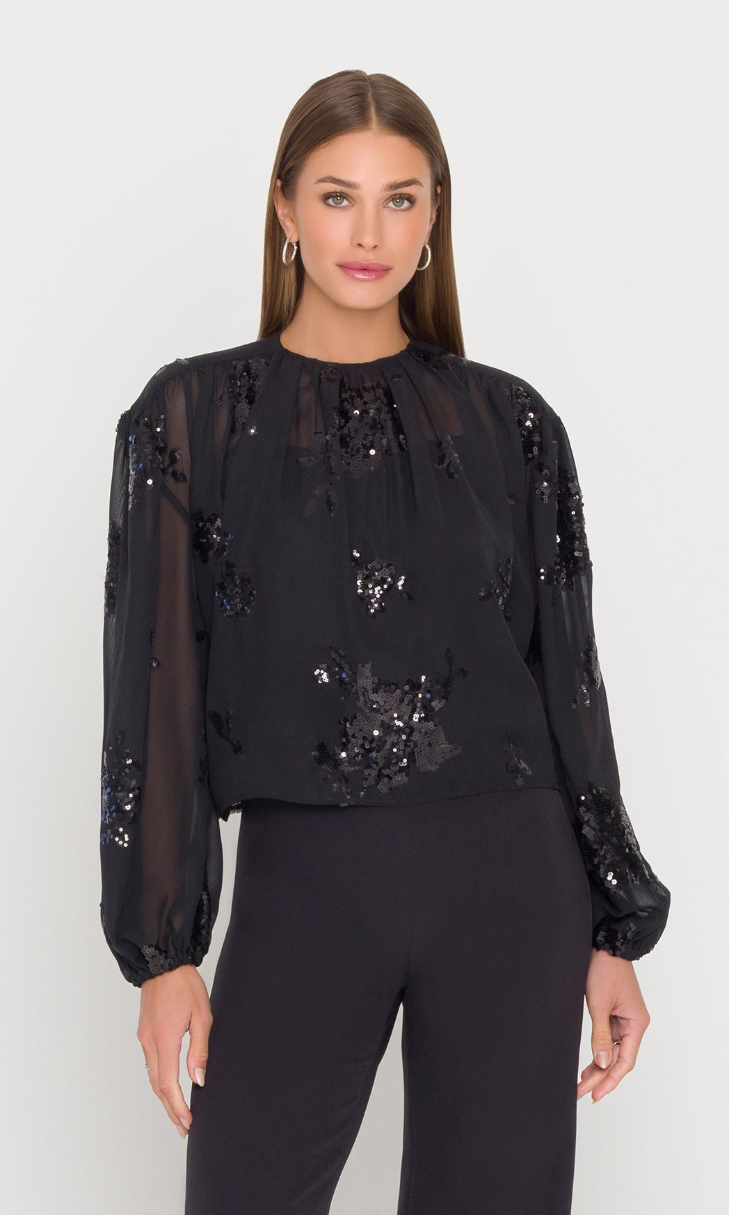 Woman wearing a black sheer top with sequins and black pants on a white background