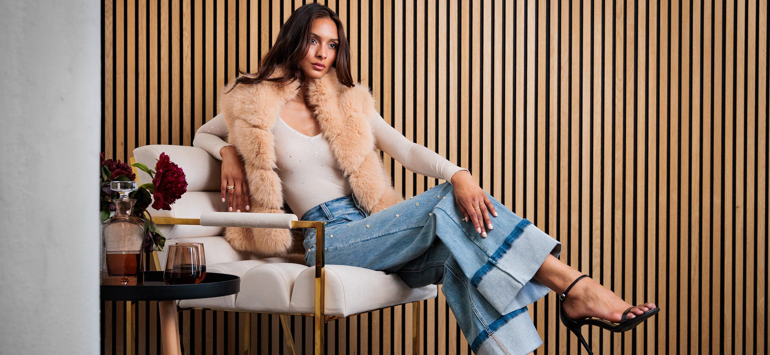 image of model sitting on a chair in front of a wooden panel wall wearing a beige faux fur vest and beige top with crystal embellished jeans and black heels