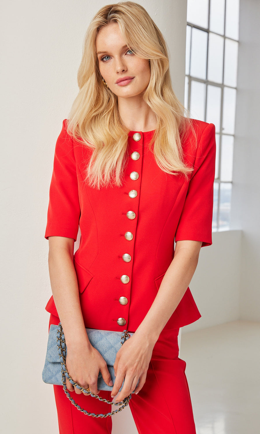 A blonde woman in a red crepe jacket with gold buttons stands by a window, striking a confident pose.