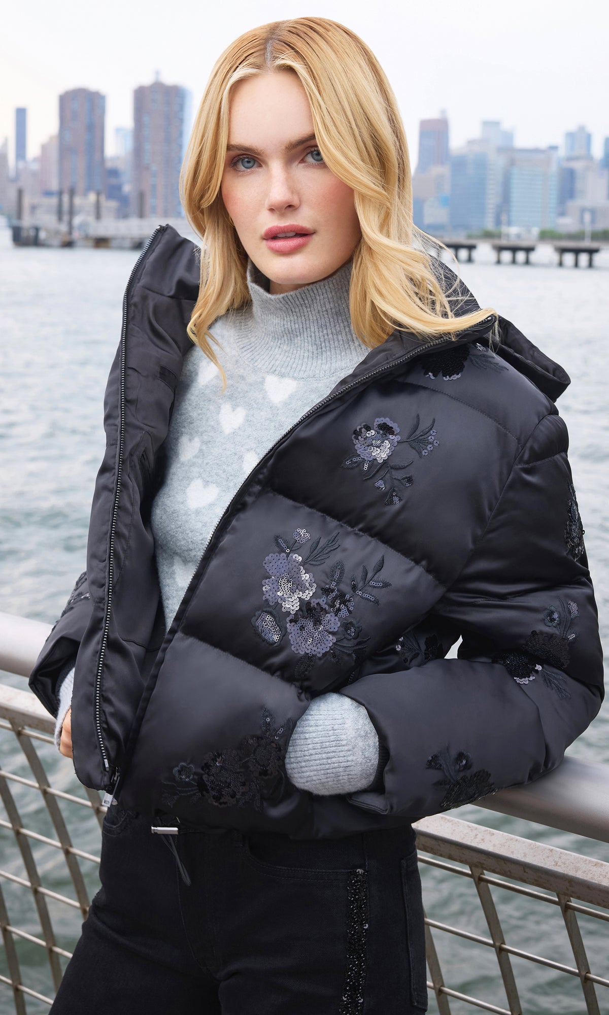 Model wearing a black puffer jacket with floral embroidery and a gray knit sweater, standing by a waterfront railing with a city skyline in the background.