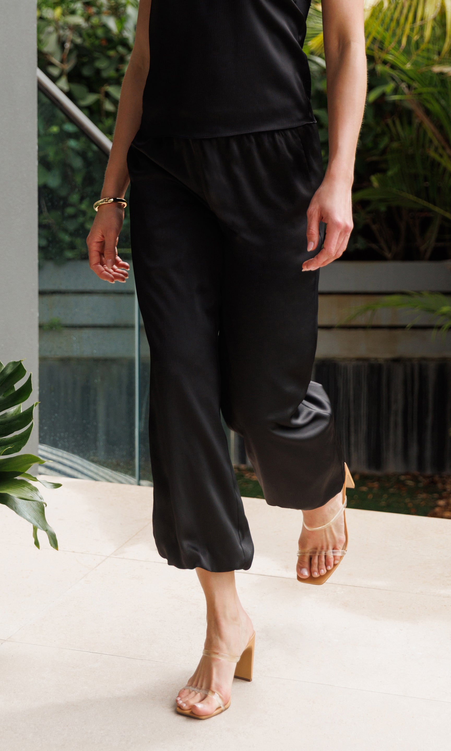 Person wearing black pants standing outdoors with greenery in the background
