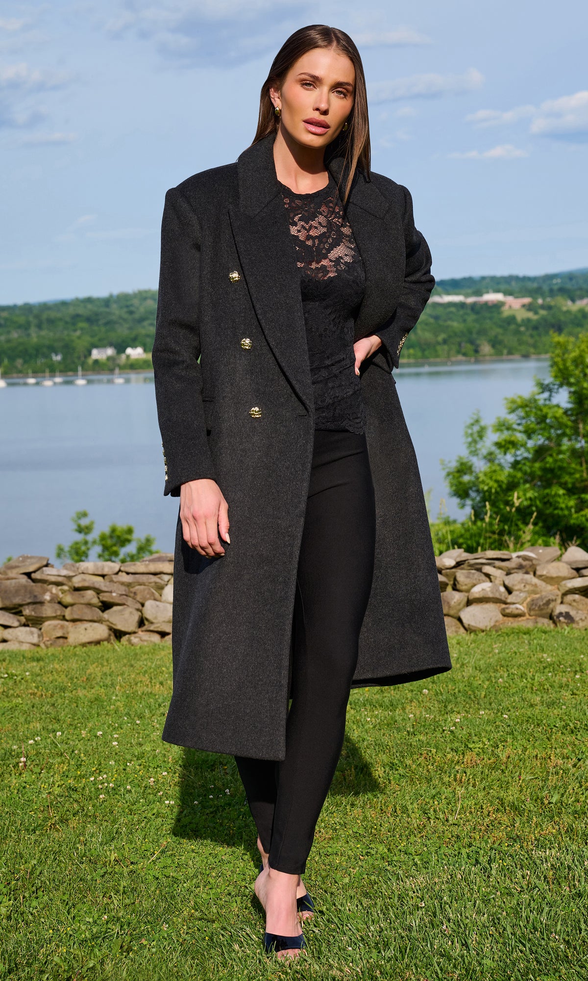 A woman stands outdoors on a grassy lawn with a lake and trees in the background, wearing a long charcoal gray double-breasted coat over a black lace top and black leggings.