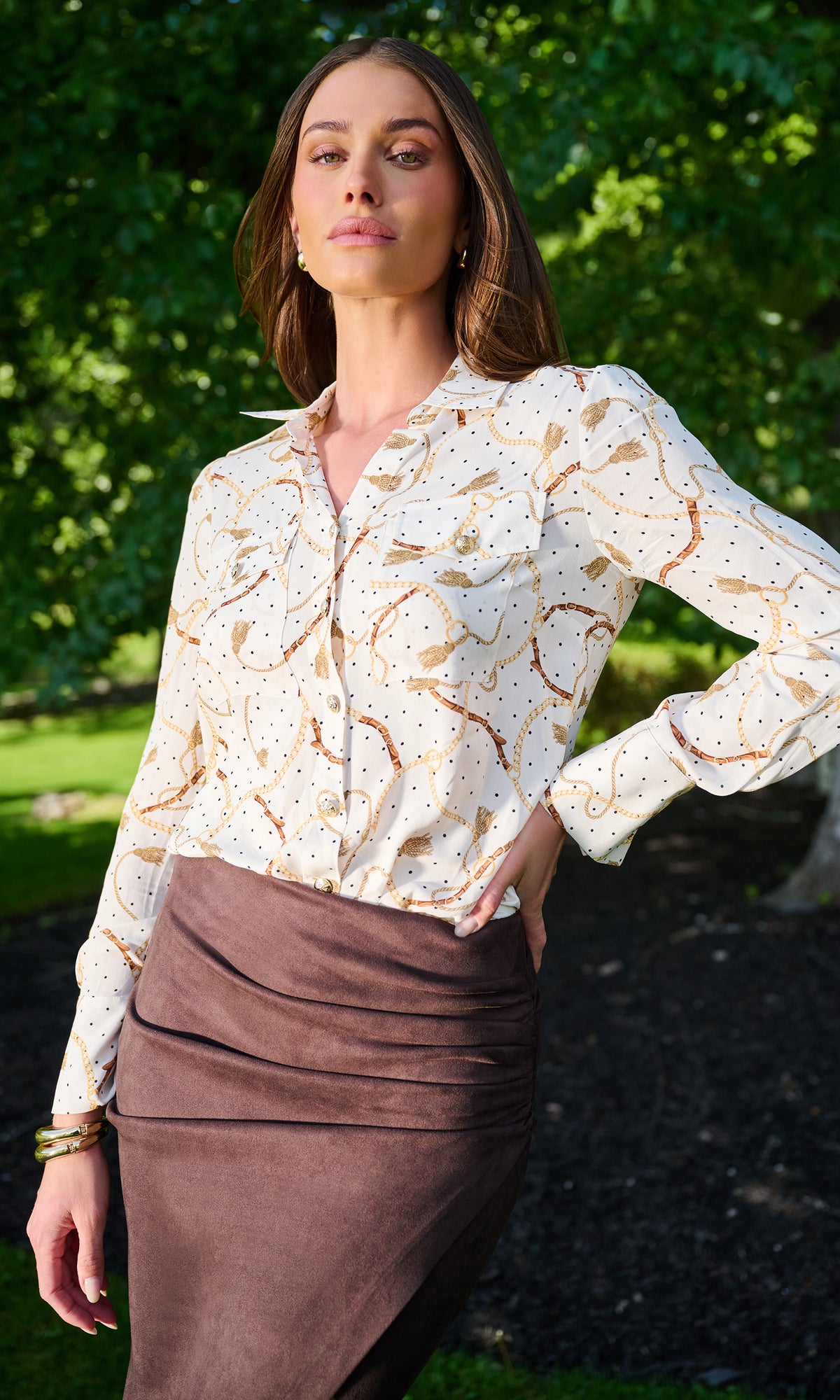 A woman wearing a white button-up blouse with a brown and gold abstract pattern, paired with a brown ruched skirt, standing outdoors in front of a green garden background.