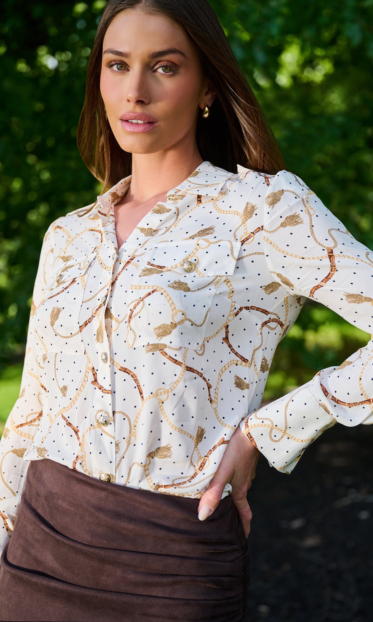 A woman wearing a white long-sleeve button-up blouse with a brown and gold abstract swirl pattern and small black polka dots, paired with a dark brown skirt.