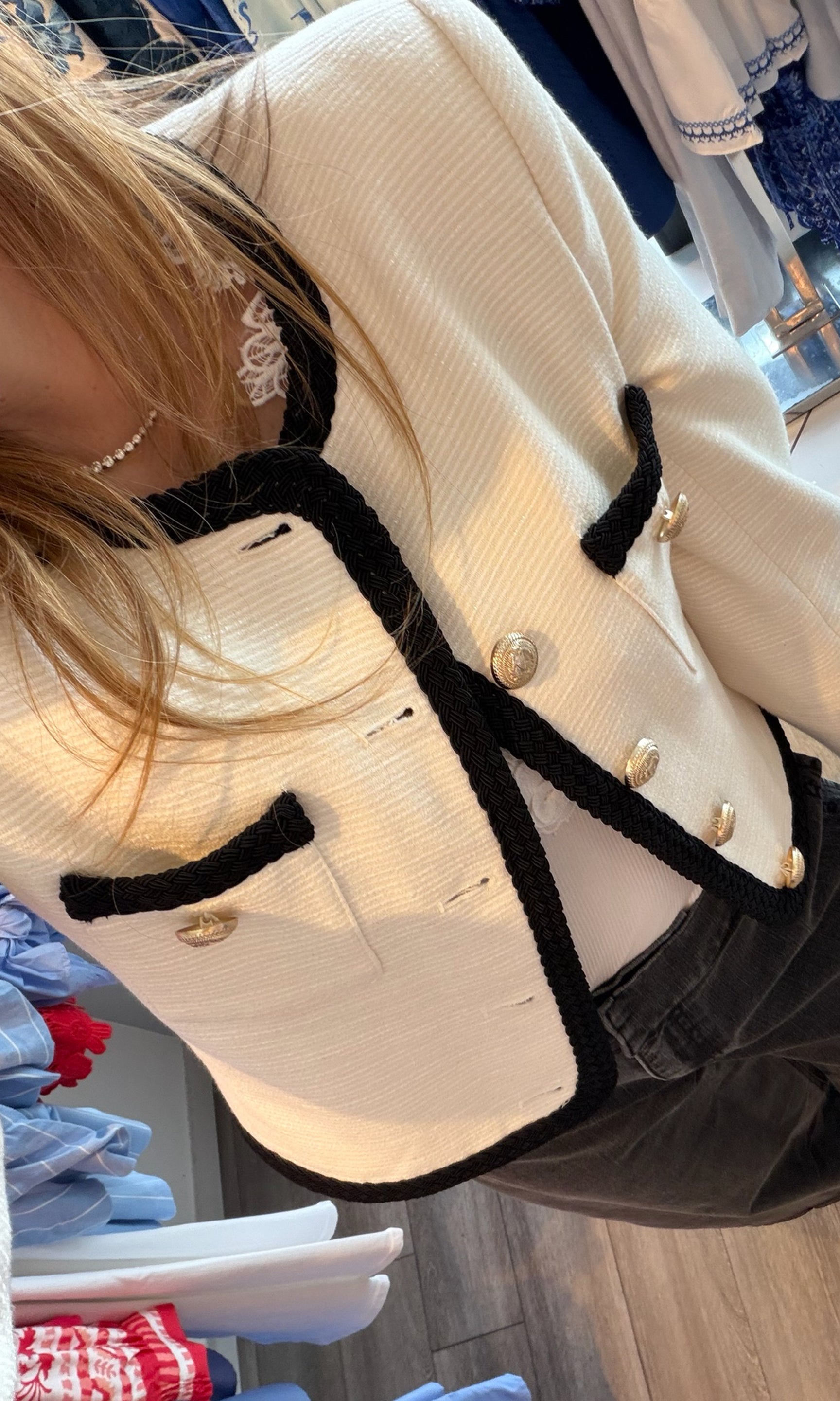Close-up of a beige coat with black trim and gold buttons, hanging on a rack.