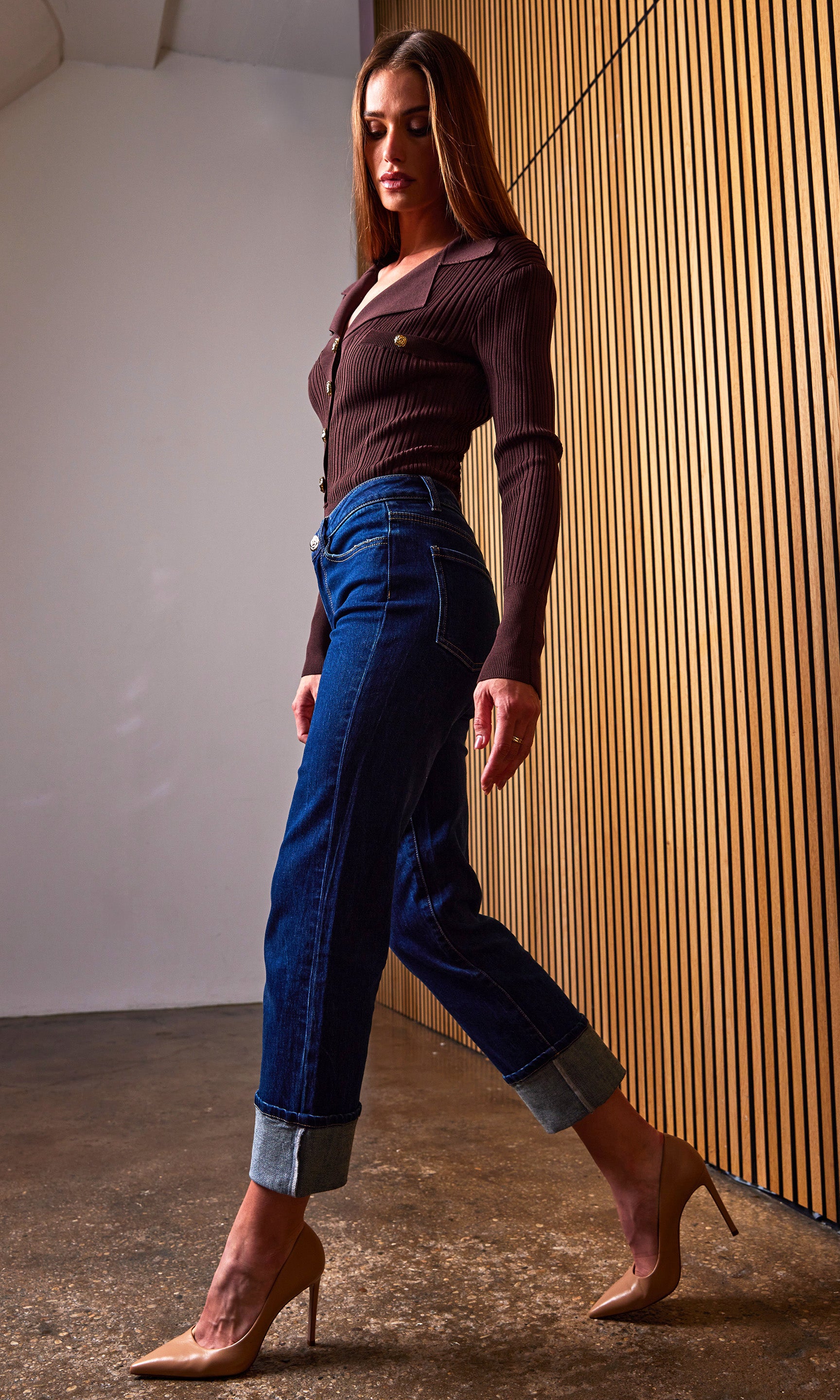 Woman in a brown top and blue jeans standing against a wooden paneled wall.