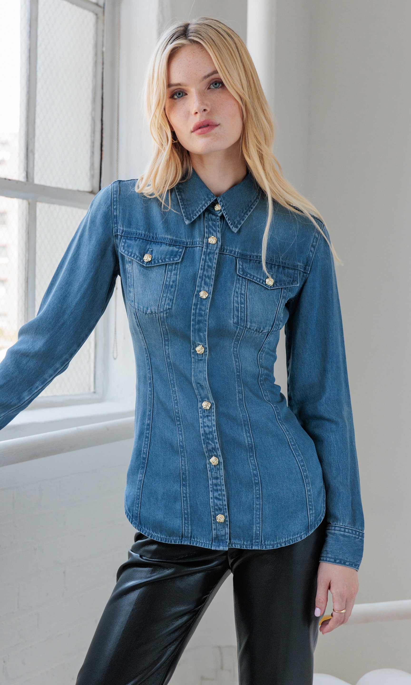 Front view of the model wearing the Antoinette Denim Shirt, styled with black leather pants and posed beside a window.