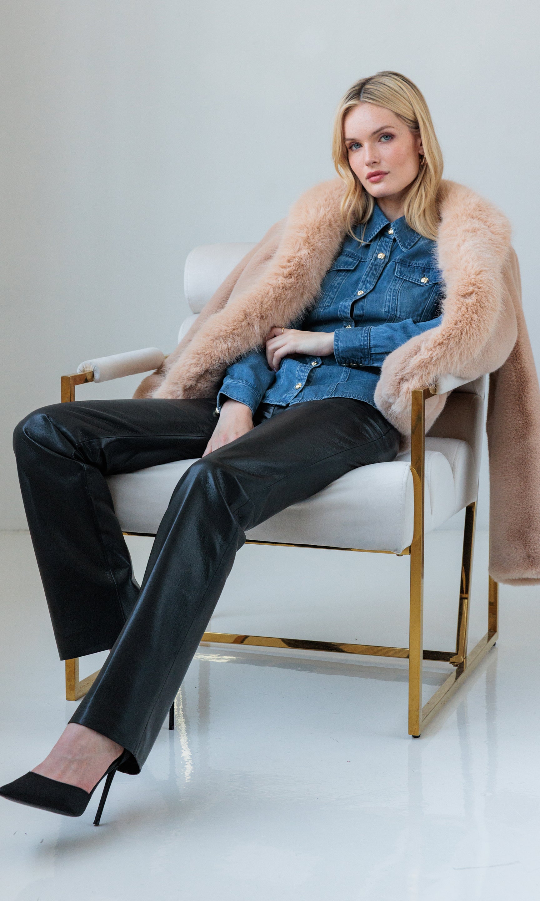 The model seated on a wooden chair in the Lynette Vegan Leather Pants and beige sweater, side angle.