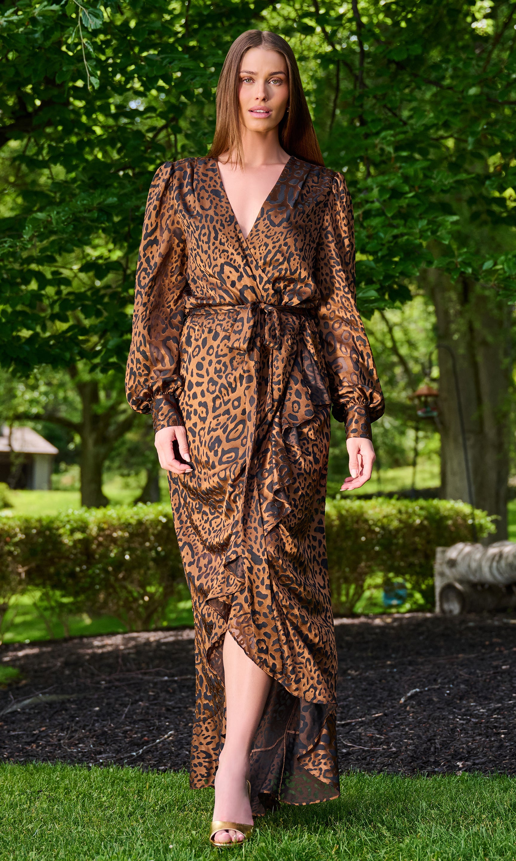 Model wearing a brown and black leopard-print maxi wrap dress with long balloon sleeves, standing outdoors on a grassy lawn.