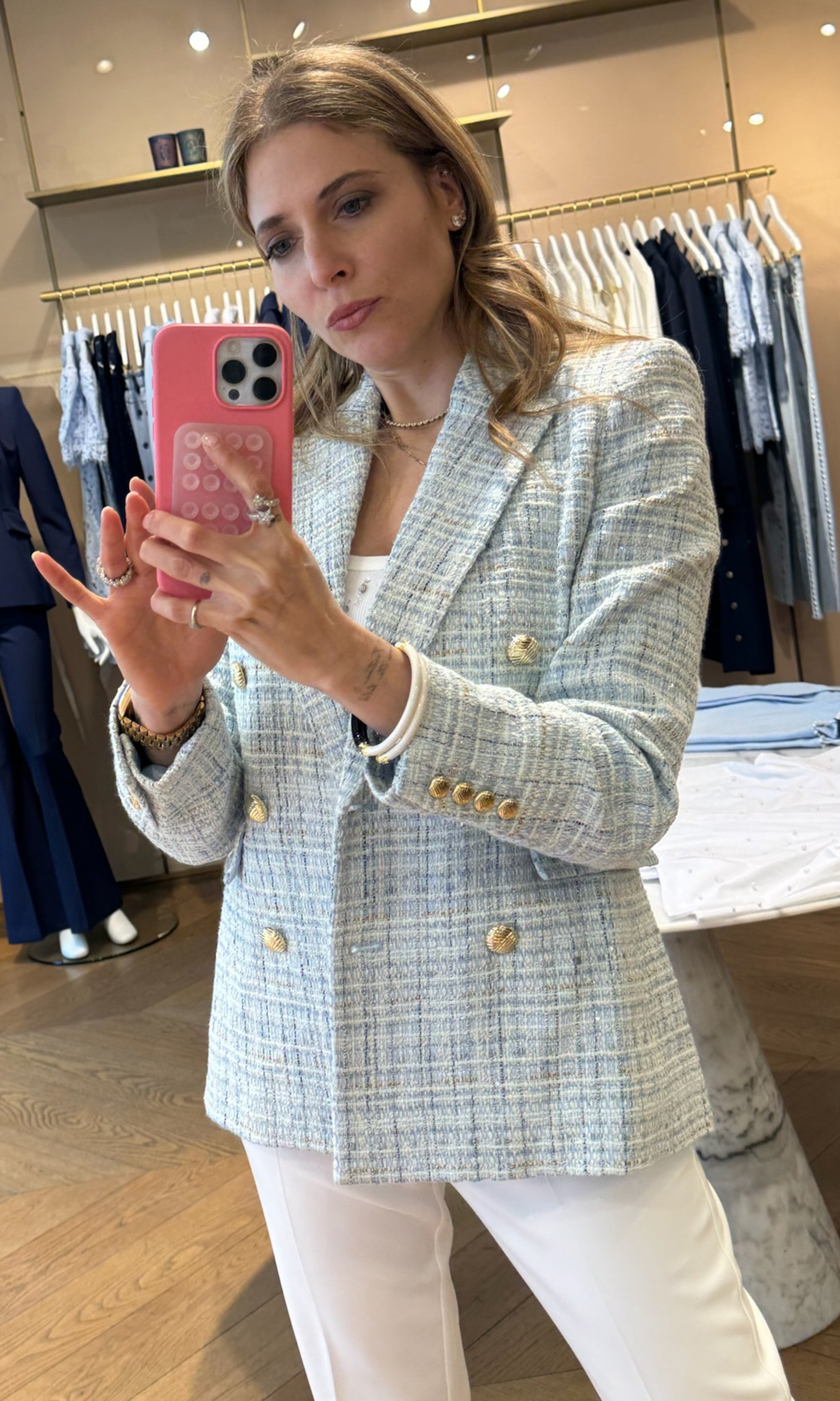 Found Roni taking a mirror selfie wearing a light-colored blazer with gold buttons in a clothing store.