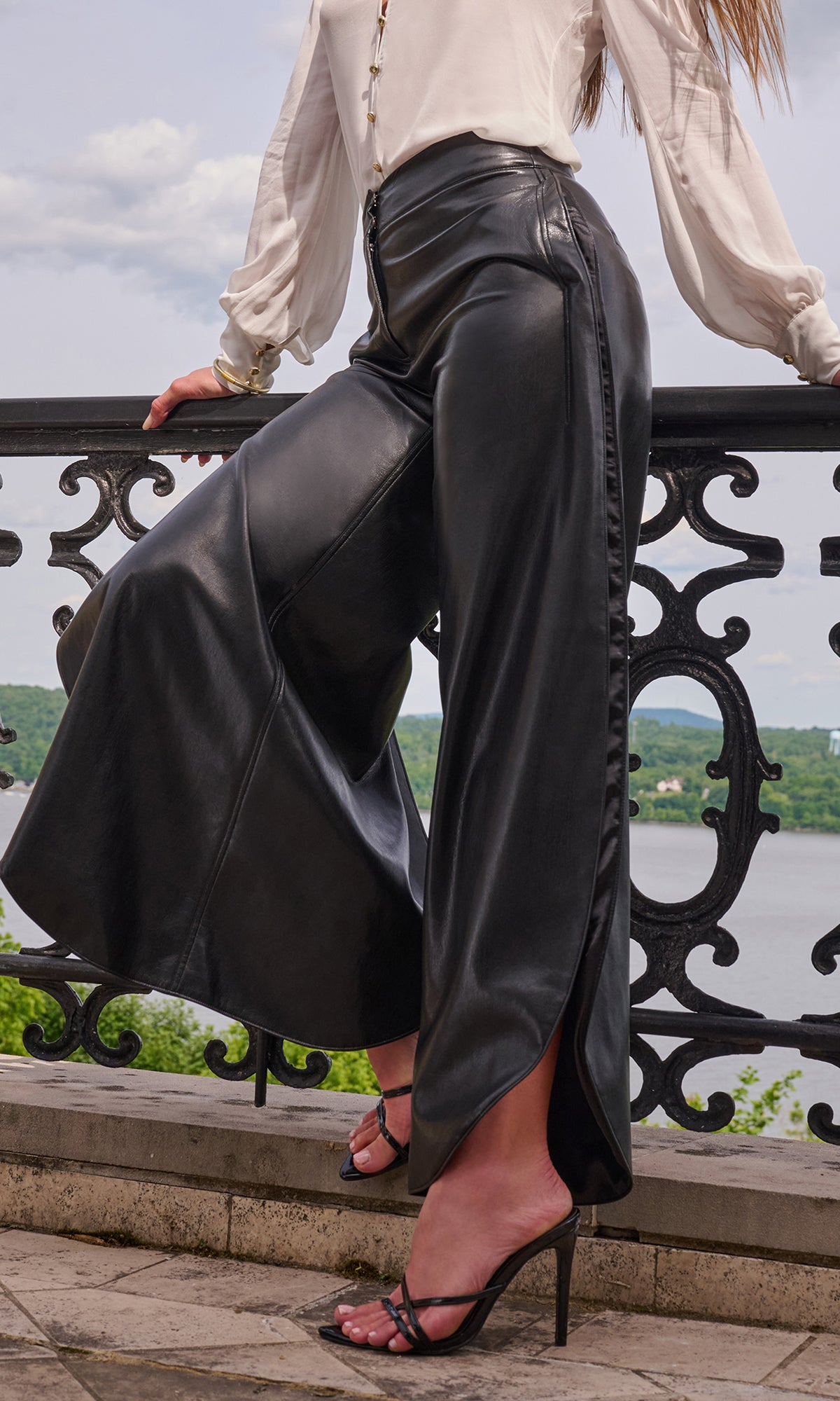 A model wearing high-waisted black faux-leather wide-leg trousers with a side slit, paired with a cream blouse and black strappy heels.
