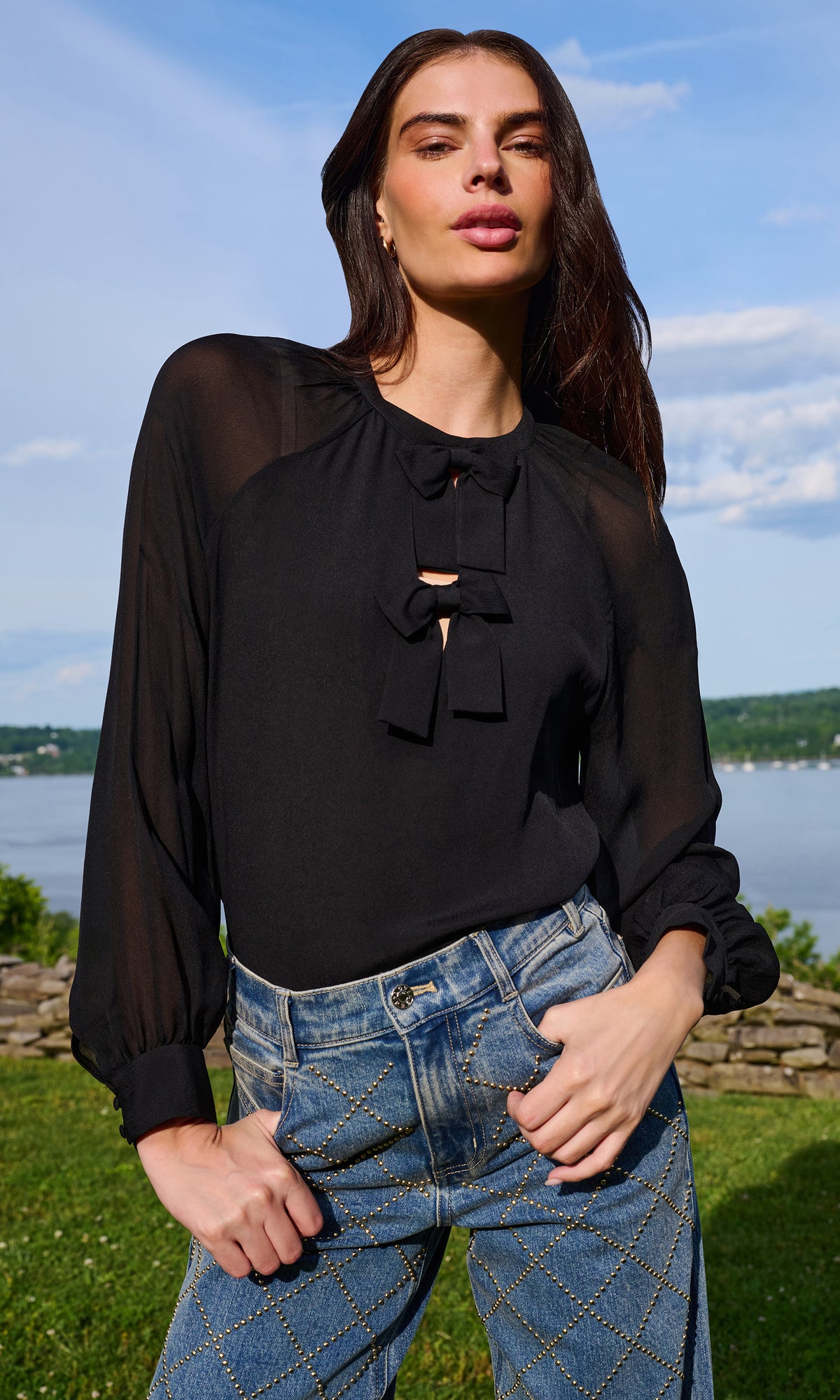 A woman wearing a sheer black long-sleeve blouse with a bow detail at the neckline, paired with blue jeans, standing outdoors with a lake and hills in the background.