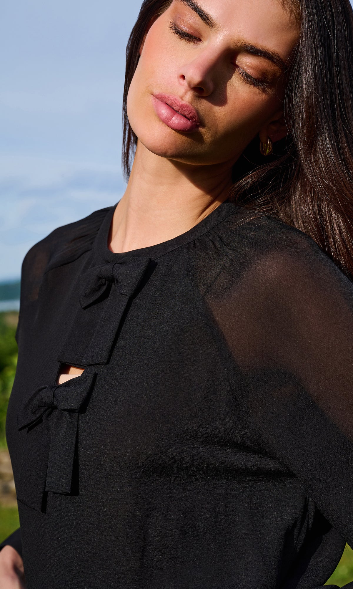 A close-up of a woman wearing a black sheer long-sleeve blouse with a bow detail at the neckline.