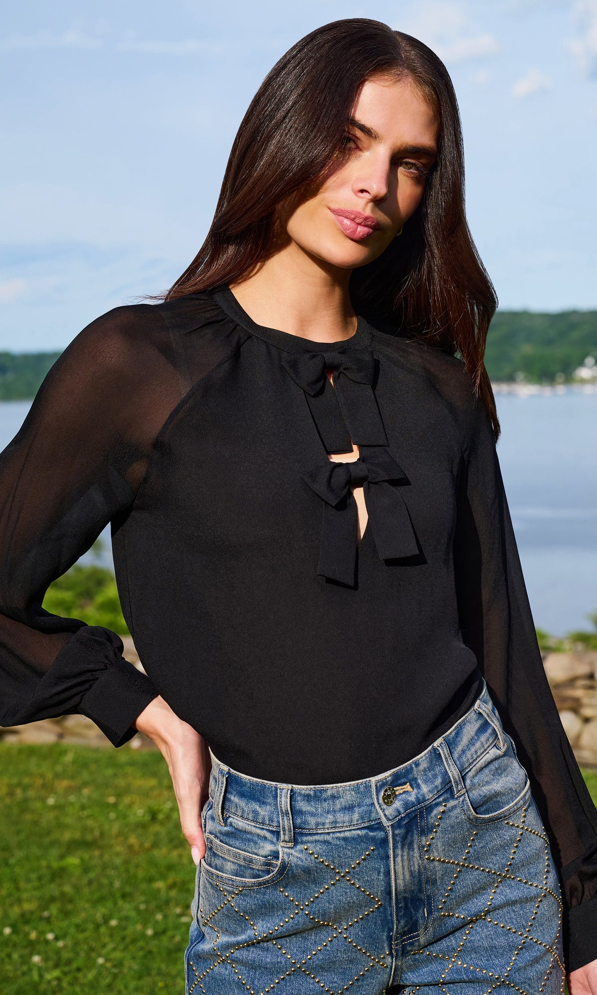 A woman wearing a black sheer long-sleeve blouse with a front bow tie detail, paired with blue jeans, photographed outdoors near water and greenery.