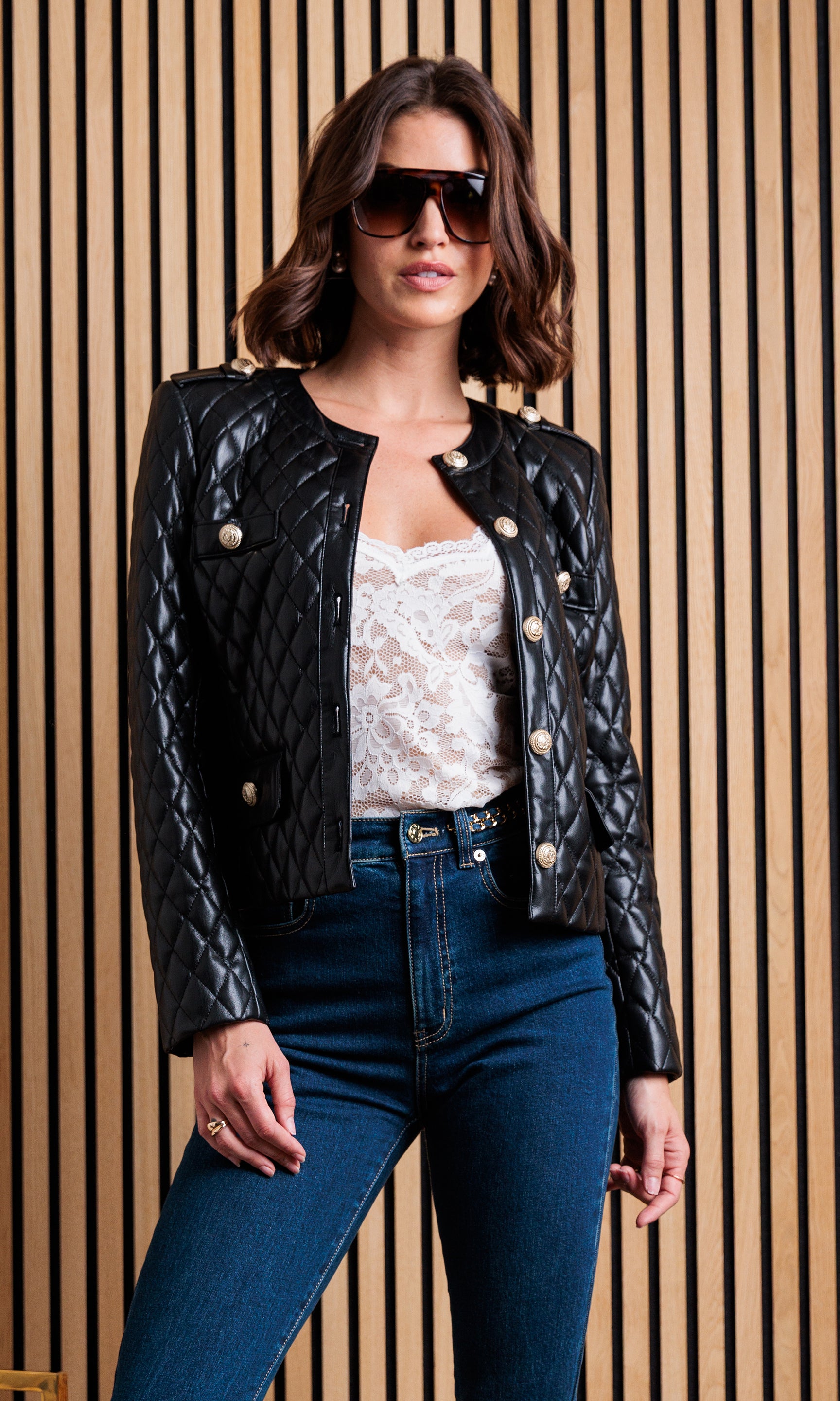 The model poses in a front view wearing the Brevel Quilted Vegan Leather Jacket in black, showcasing its quilted texture against a wood-paneled backdrop.