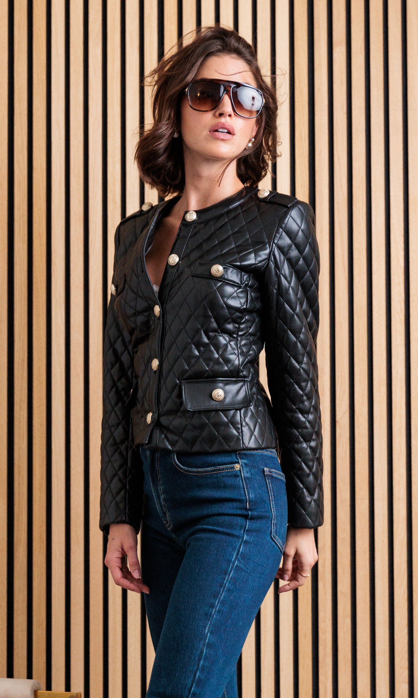 The model appears in a three-quarter side view wearing the Brevel Quilted Vegan Leather Jacket in black, highlighting its tailored fit and gold button details.