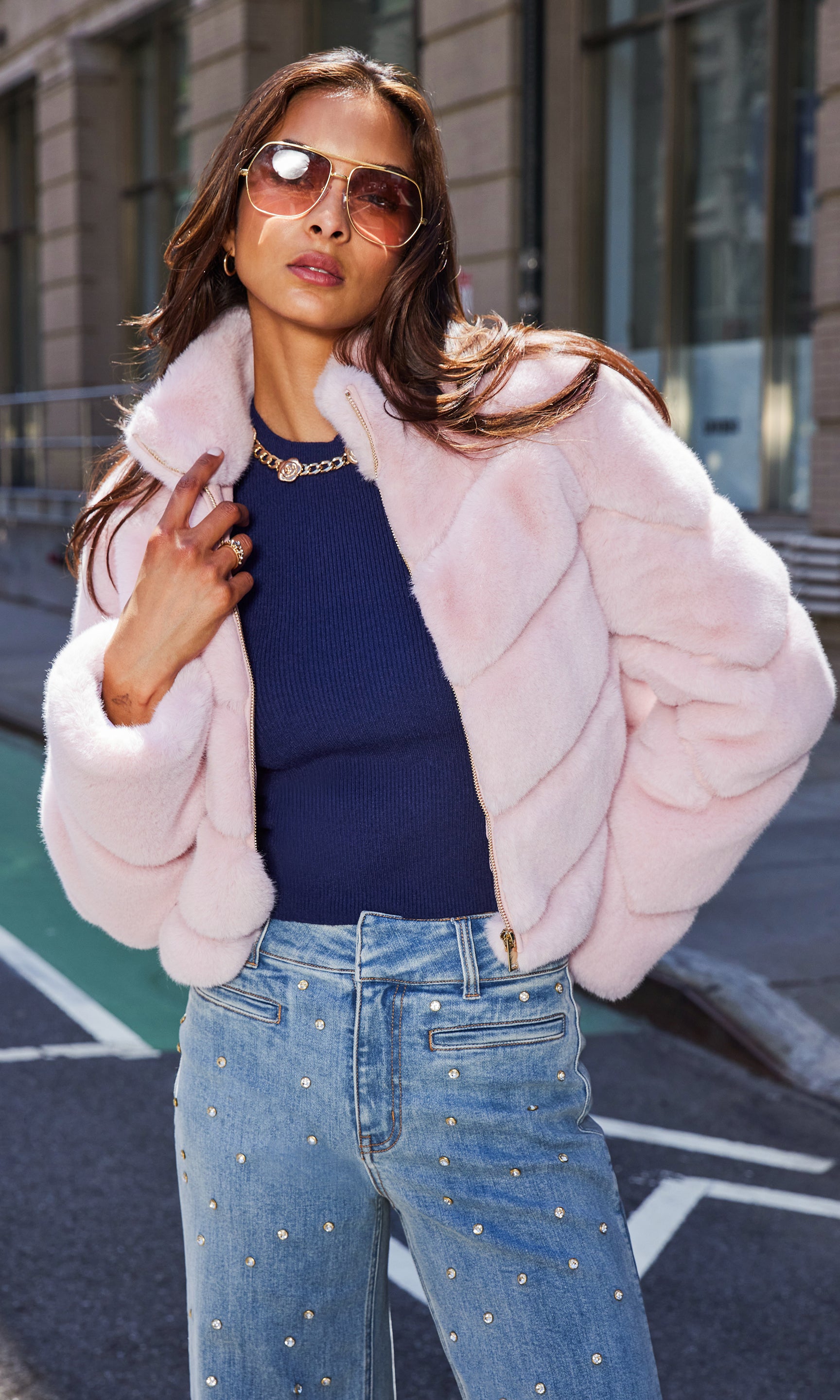The model appears in a front view wearing the Brianna Faux Fur Jacket in ballet slipper layered over a navy sweater and high-waisted, pearl-embellished jeans, turning her head to showcase the jacket’s luxurious finish and structured shoulders.