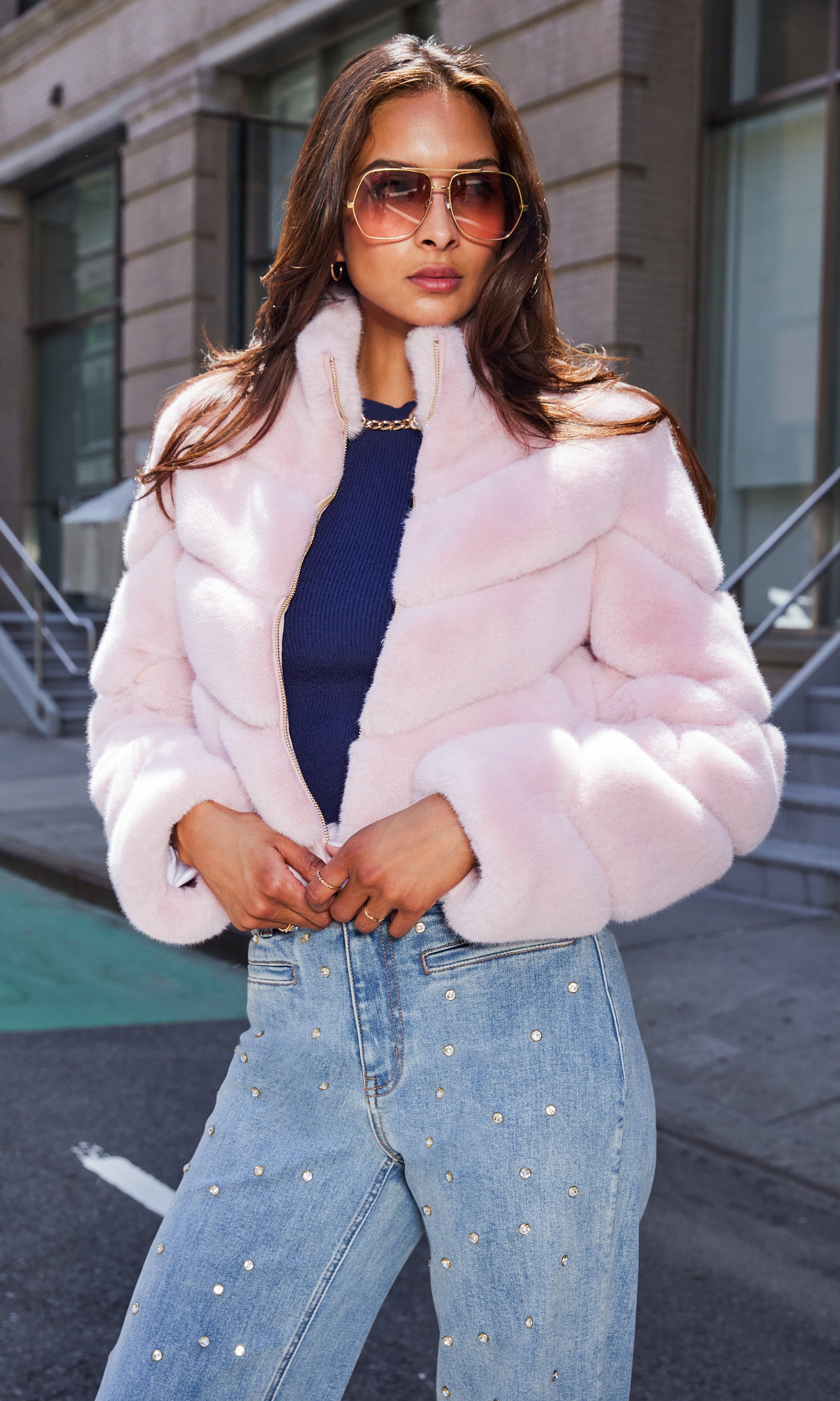 The model poses in a front view wearing the Brianna Faux Fur Jacket in ballet slipper layered over a navy sweater and high-waisted, pearl-embellished jeans, showcasing its plush chevron quilting and high collar against an urban backdrop.