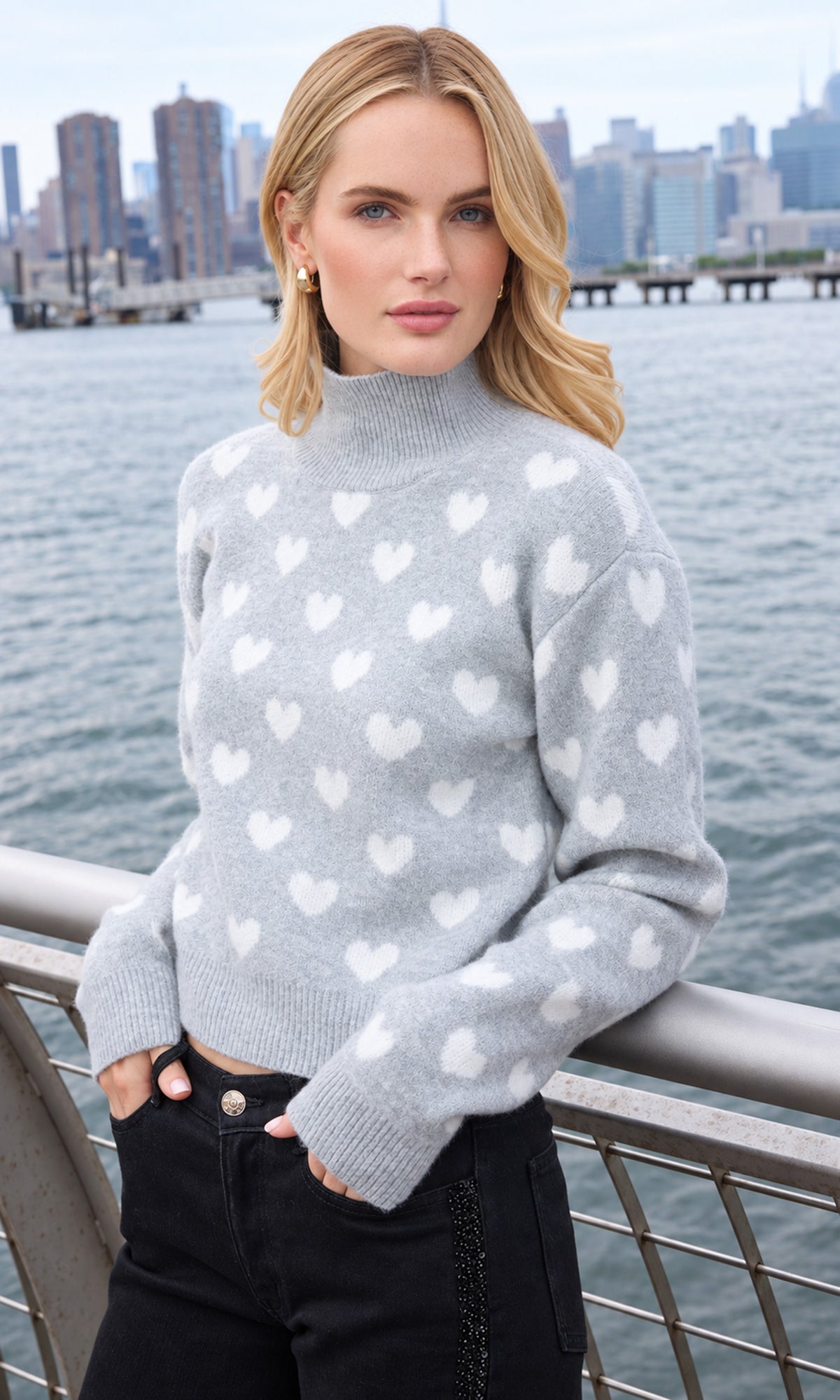 A woman wearing a light gray mock-neck knit sweater with a white heart pattern, paired with black pants, standing outdoors with a city skyline and water in the background.