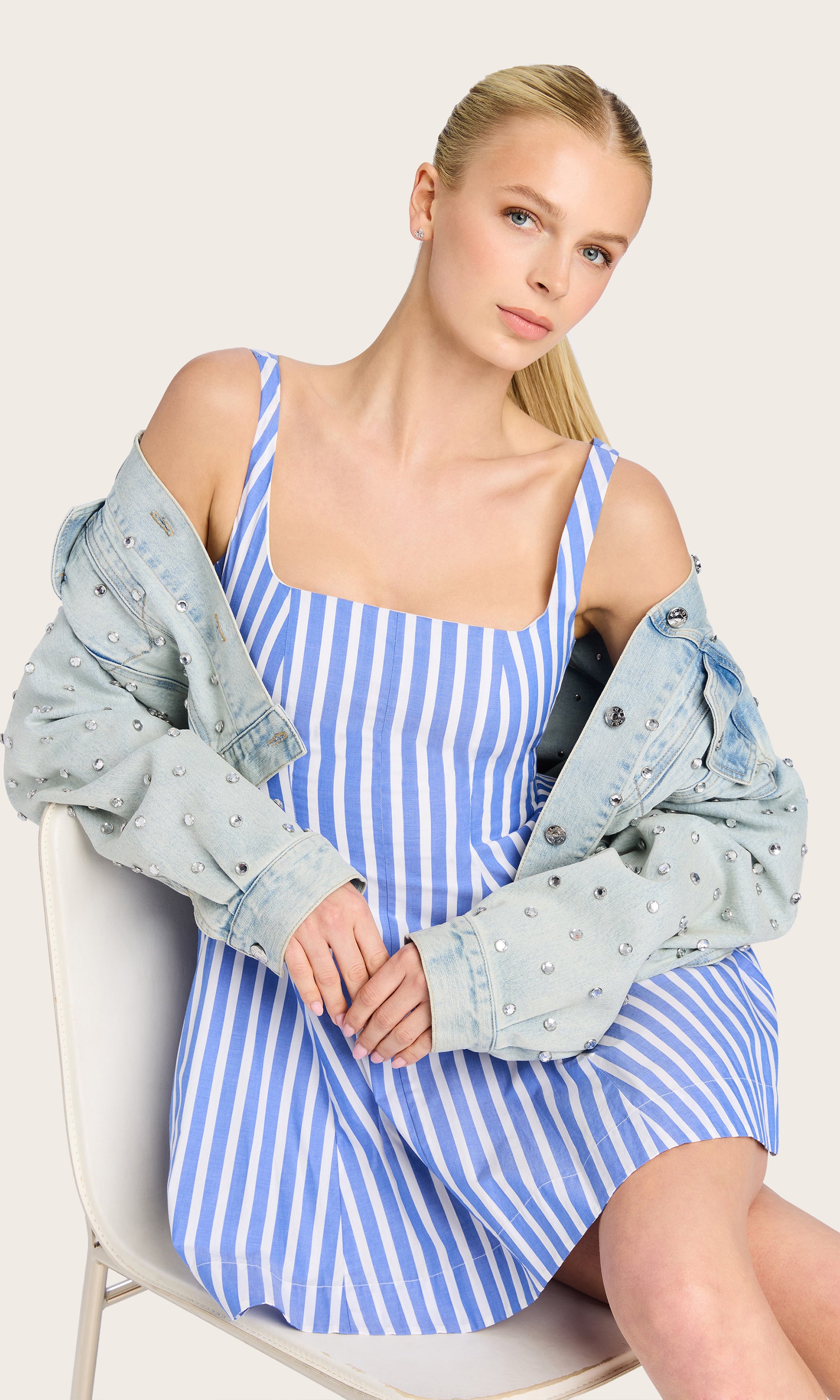 Inmage of model sitting on a chair wearing the Courtney mini dress in a white/blue striped pattern with her hair in a ponytail and a blue denim jacket with crystal embellishments all over.