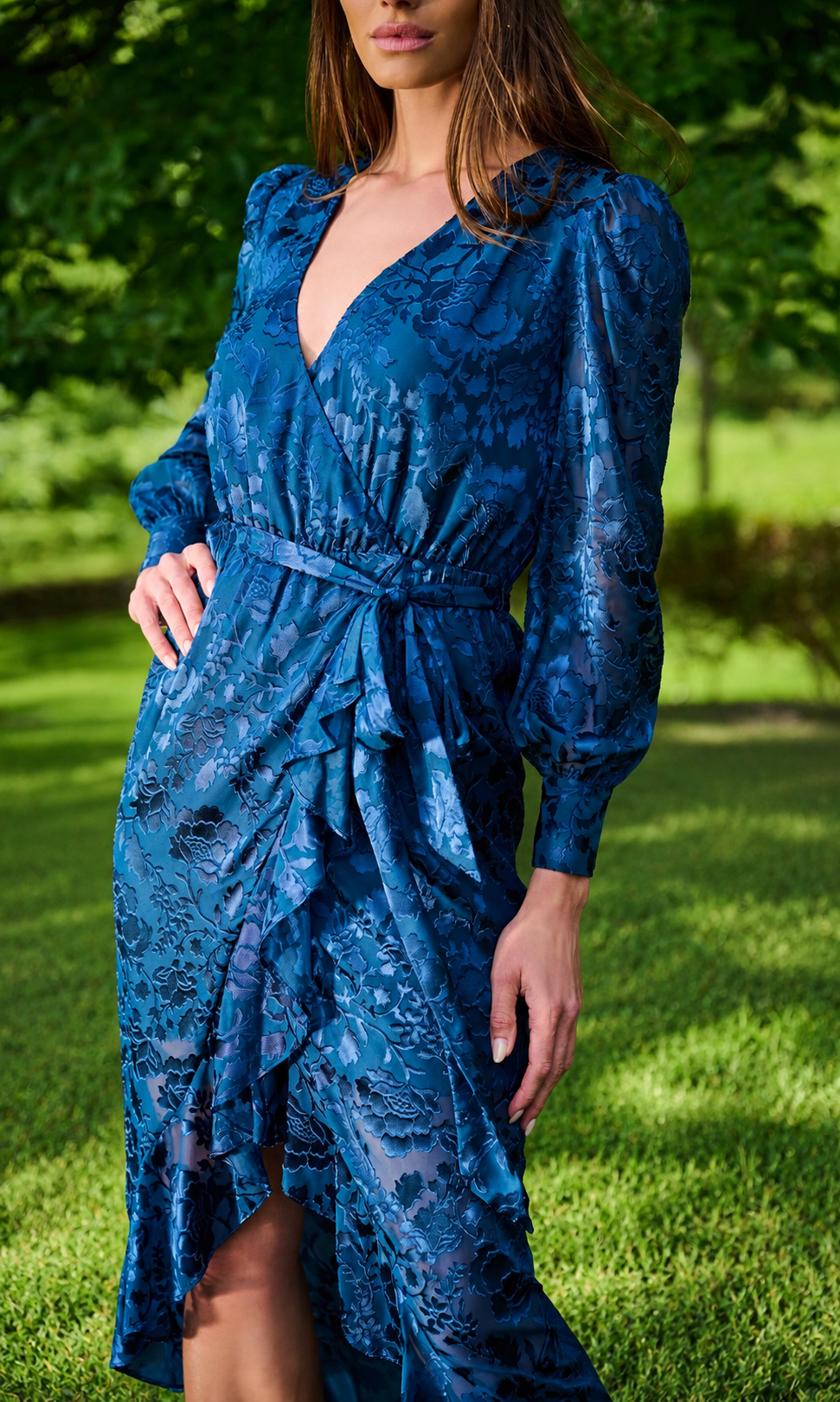 Model wearing a blue patterned midi dress with long puff sleeves, and a matching waist tie, photographed outdoors in a park setting.