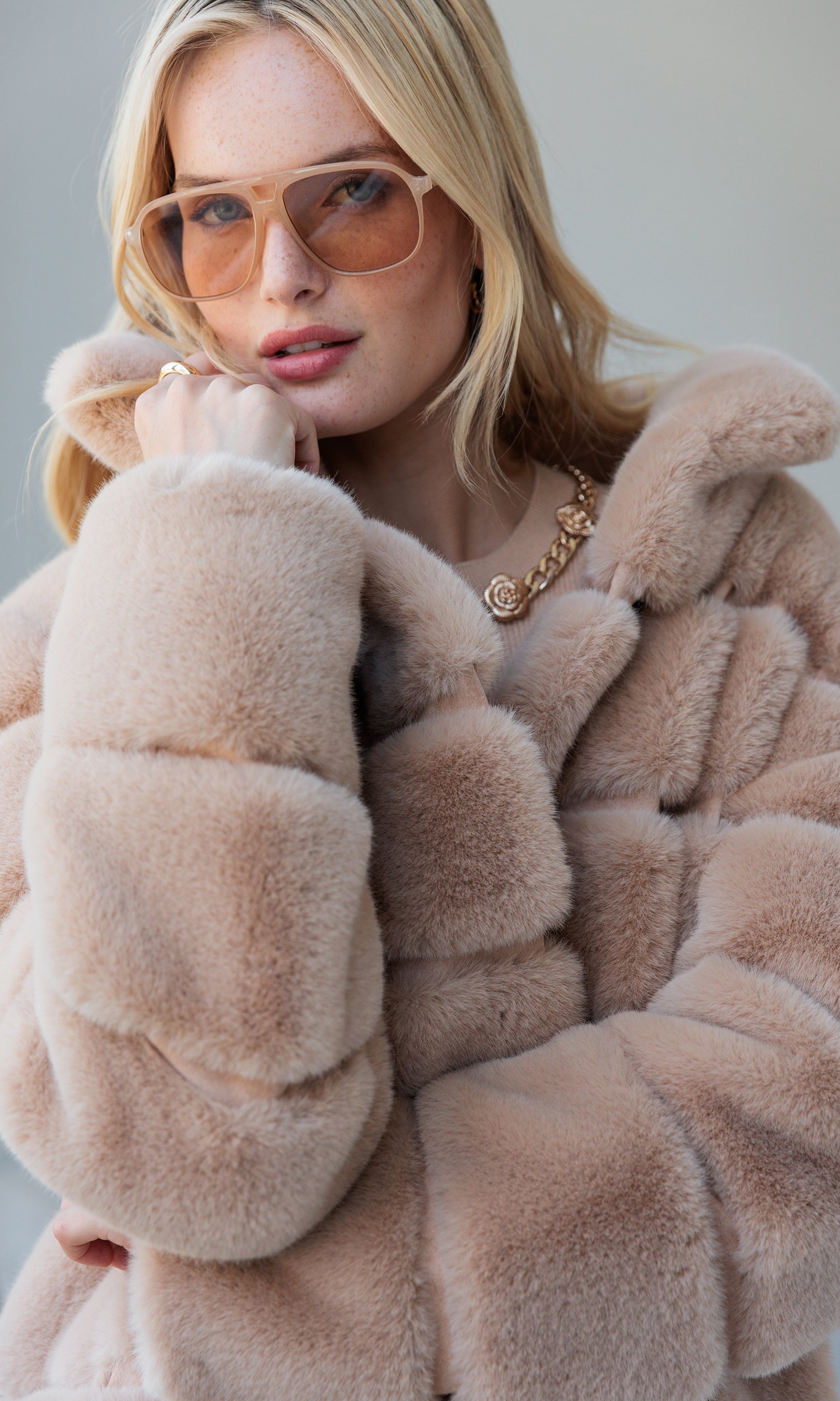 Woman wearing a beige fur coat and sunglasses with a neutral background