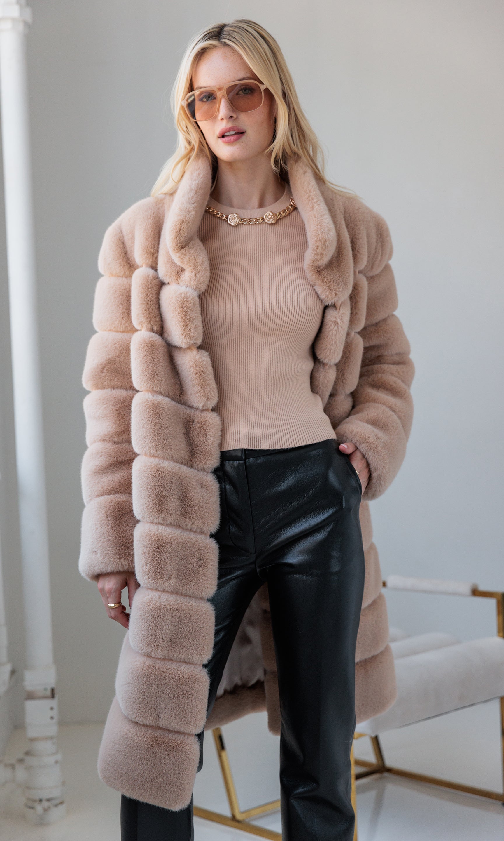 Woman wearing a long fur coat, beige top, and black faux leather pants indoors.
