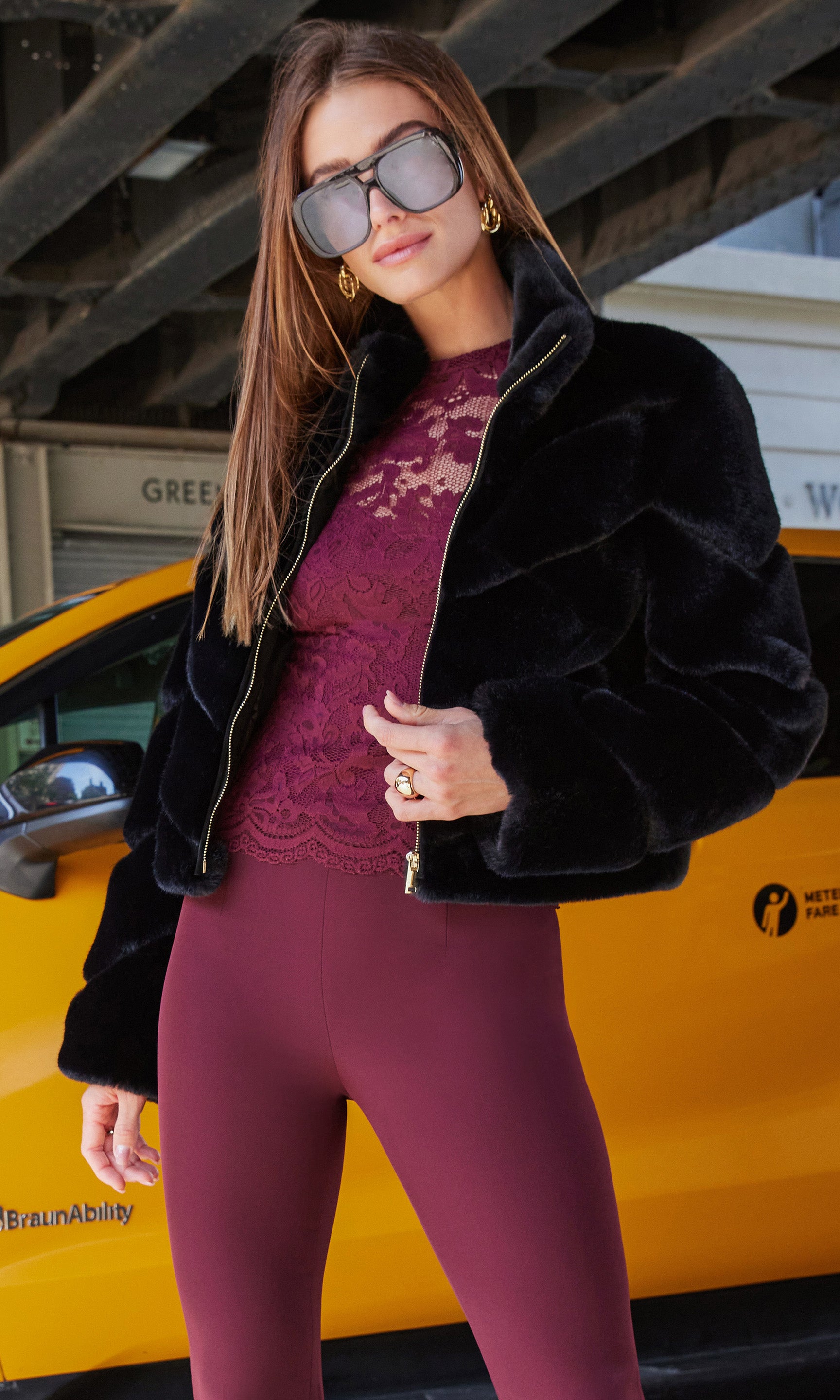 The model poses in a front view wearing the Brianna Faux Fur Jacket, highlighting the jacket's plush black texture paired with a burgundy lace top and matching pants.