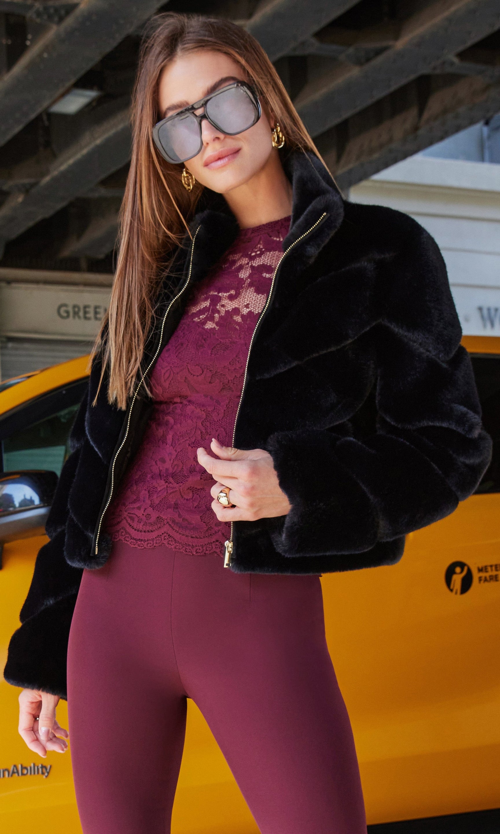 Woman in a stylish outfit with a black fur jacket and burgundy top and pants standing in front of a yellow taxi.