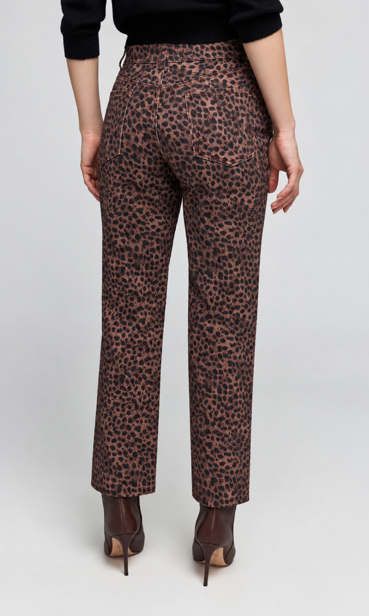 A person shown from the back wearing leopard-print trousers, a black long-sleeve top, and brown pointed-toe heels against a light gray studio background.