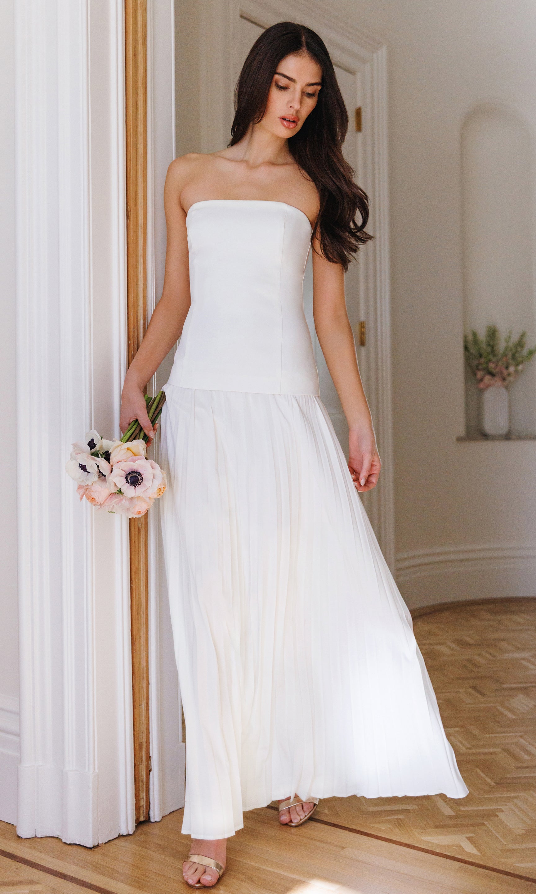 Woman in a strapless white flowy dress holding flowers indoors.