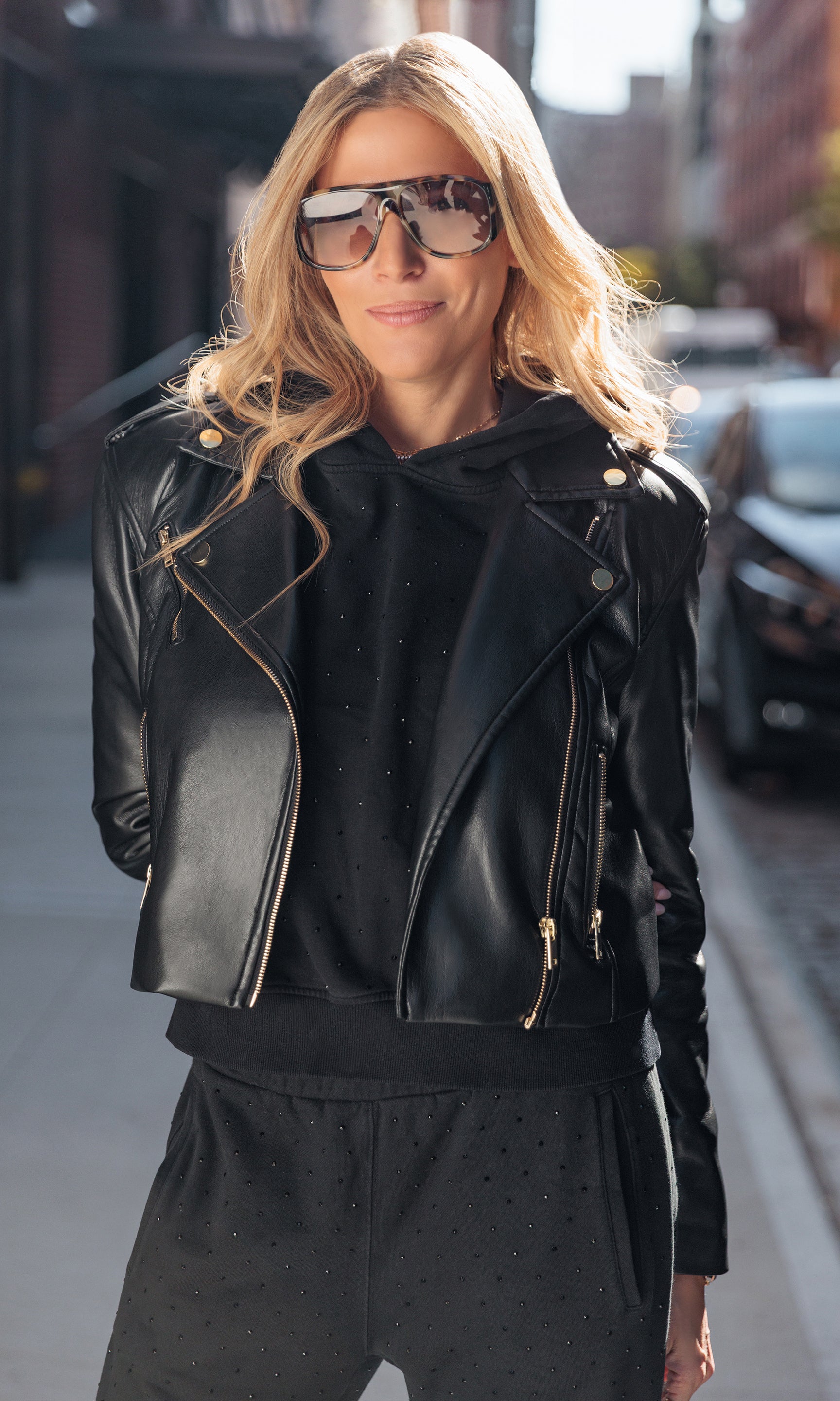 Woman wearing a black leather jacket and sunglasses on a city street.