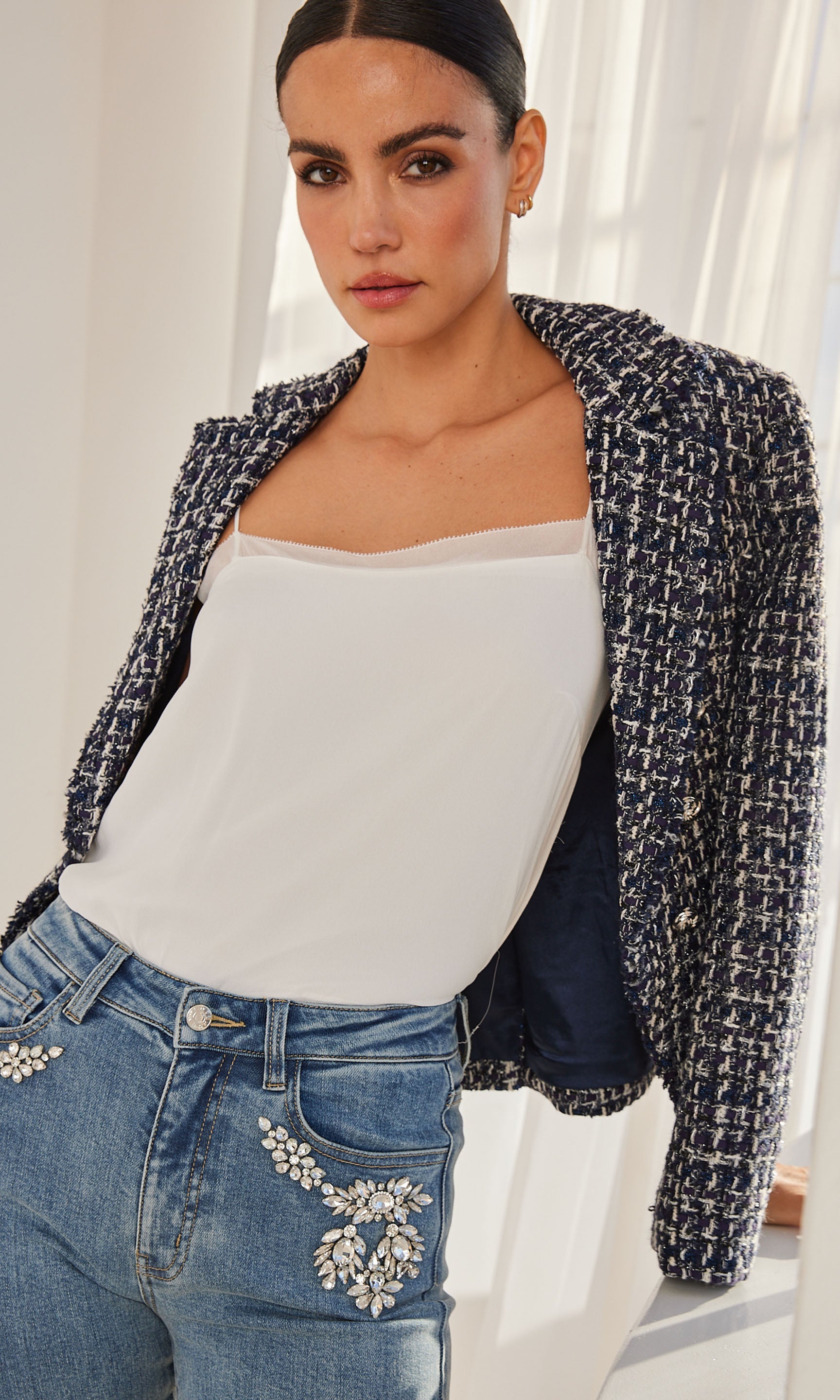 Close-up of a model wearing a white cami with thin straps and delicate detailing along the neckline, paired with a textured navy tweed blazer.