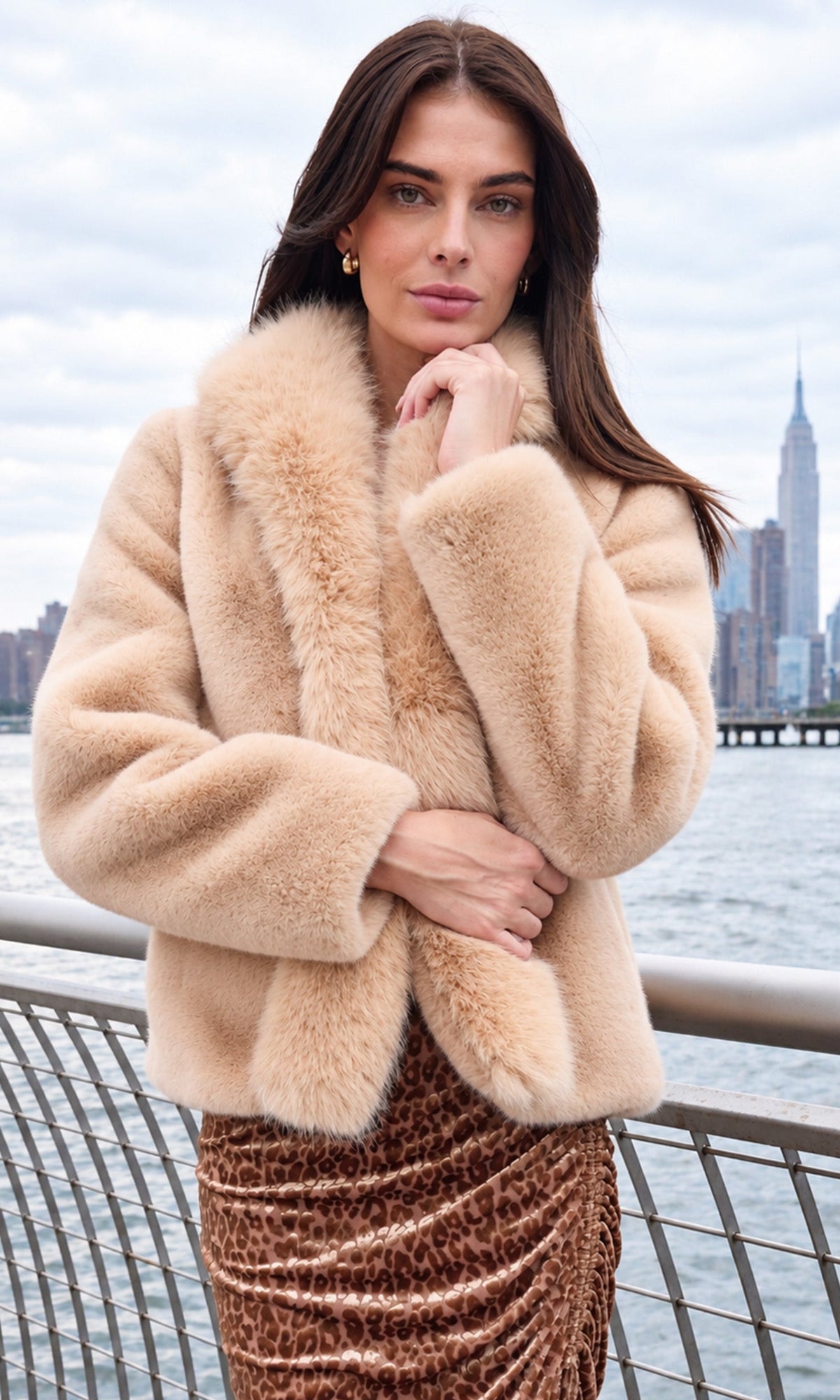 Model wearing a beige faux fur jacket with a leopard print skirt, standing in front of a waterfront with a city skyline in the background.