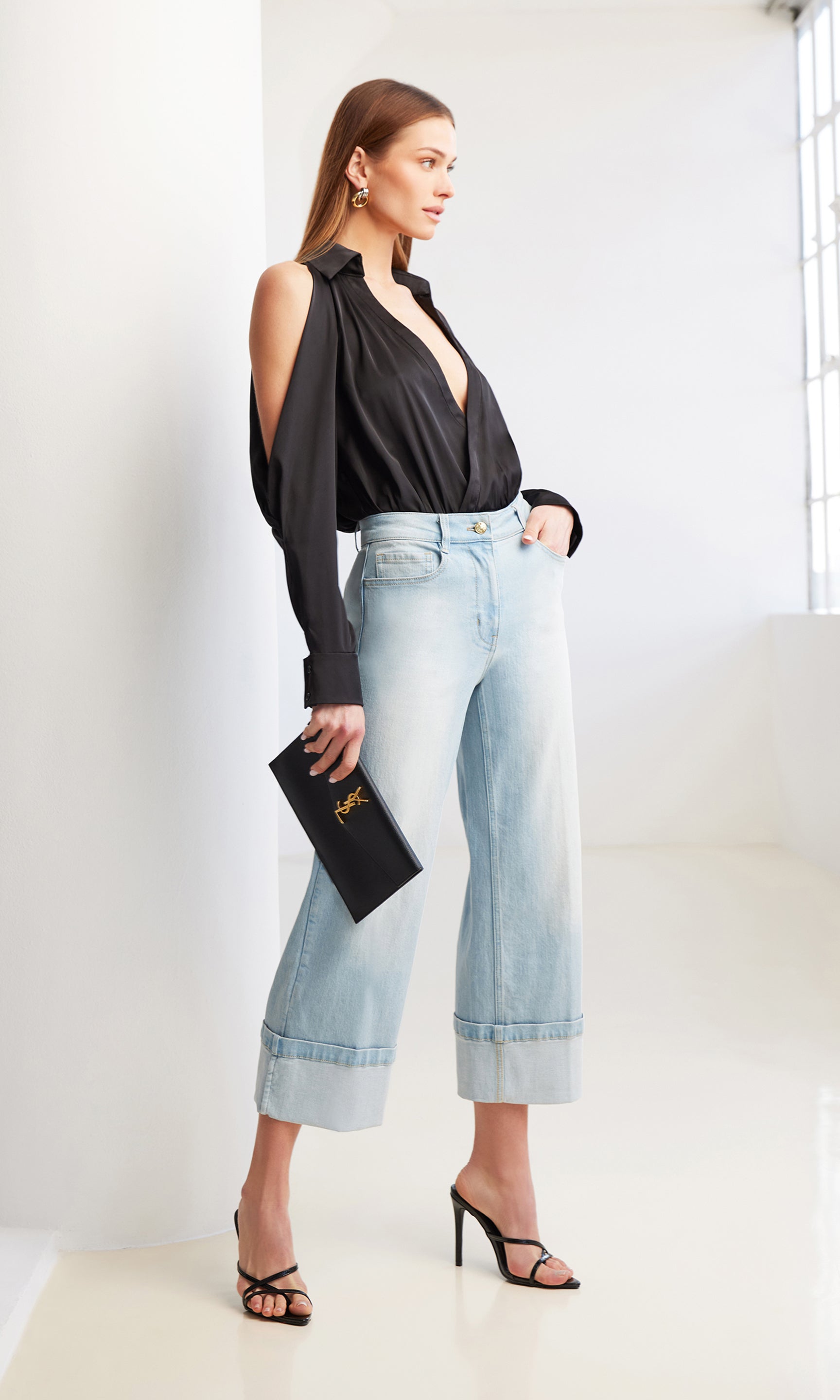 image of model posing in front of a window wearing a black bodysuit, paired with the Eros jeans in blue, holding a black clutch purse, paired with black heels