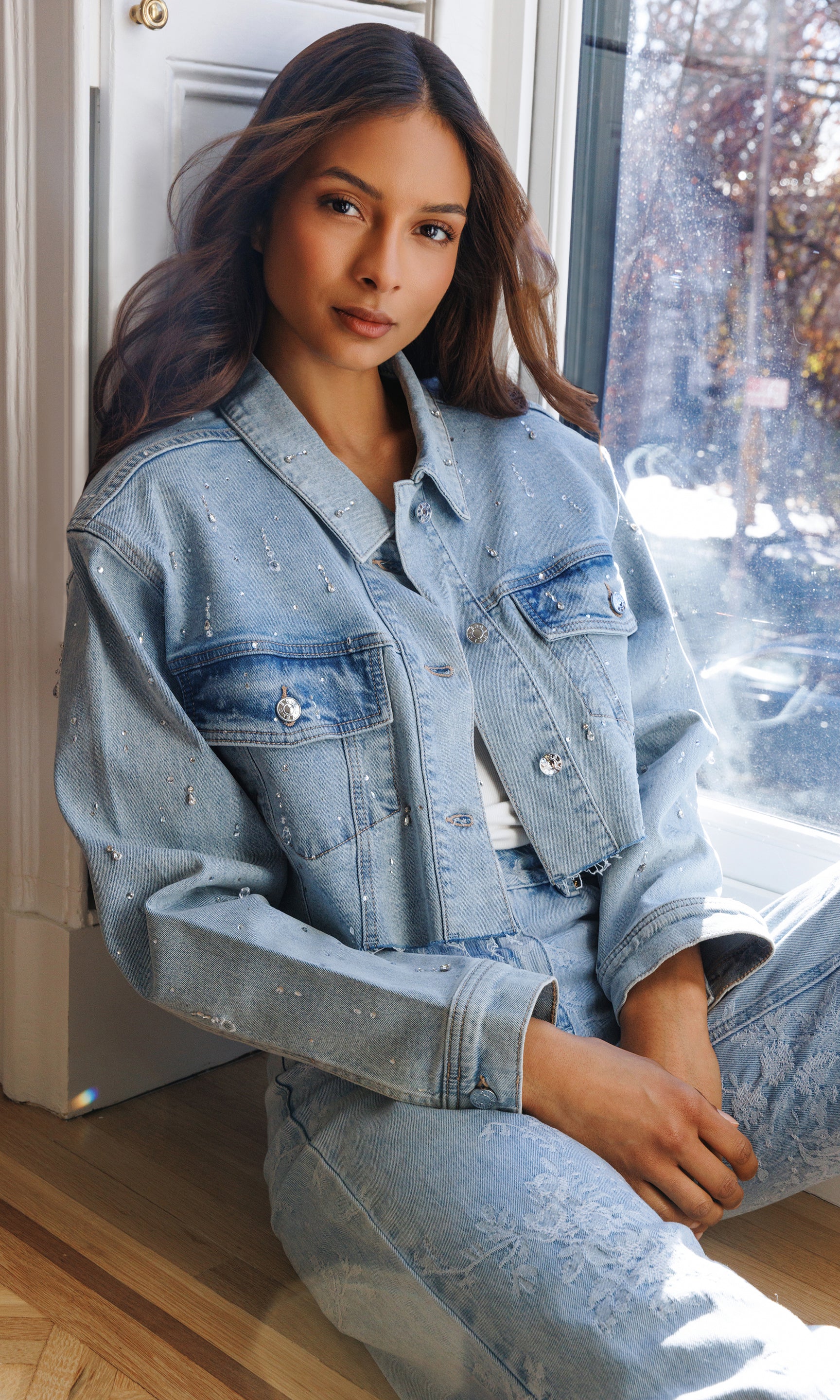 Woman wearing a light blue denim jacket and pants sitting by a window.