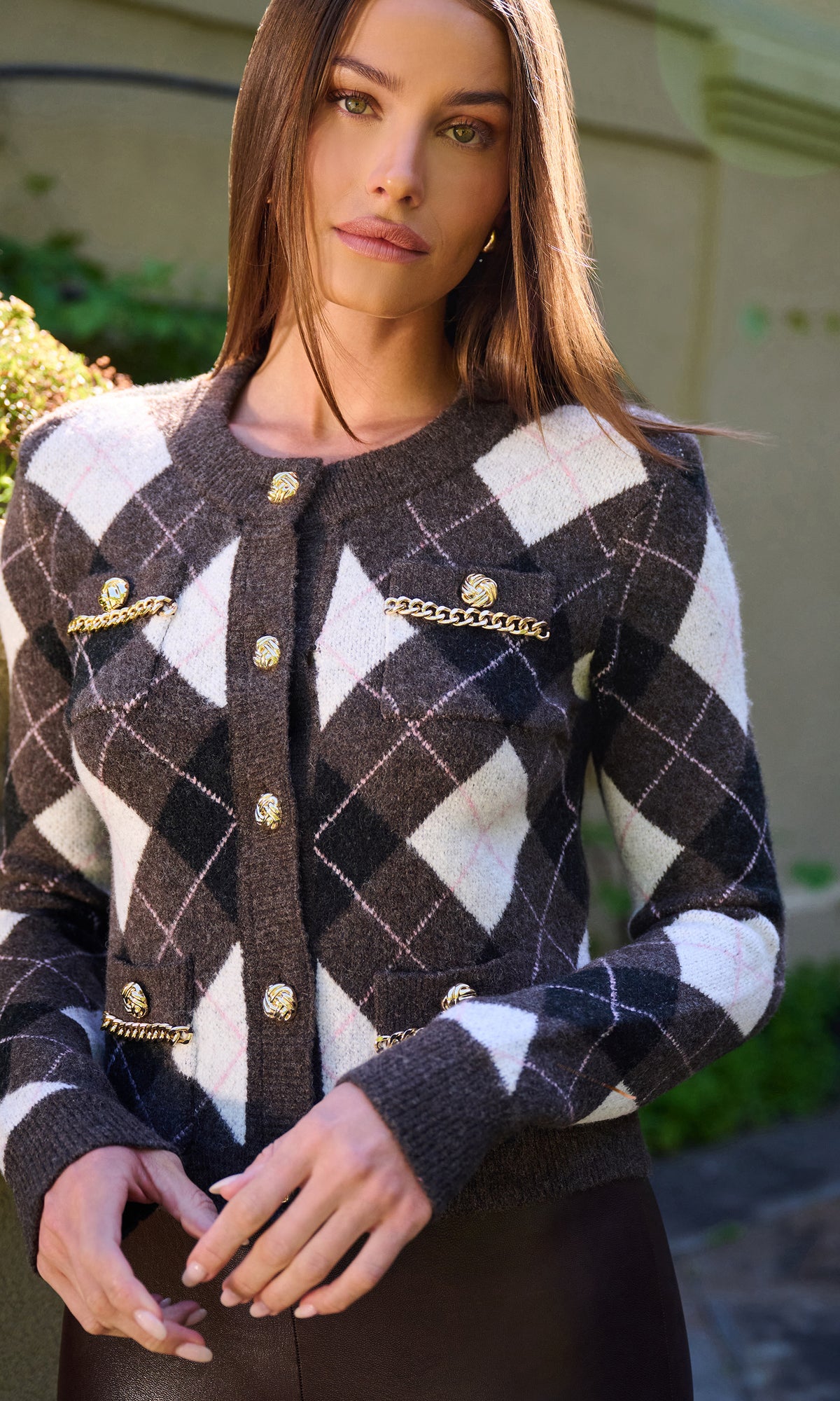 A woman wearing a dark brown and white argyle knit cardigan with gold-tone chain and button details.