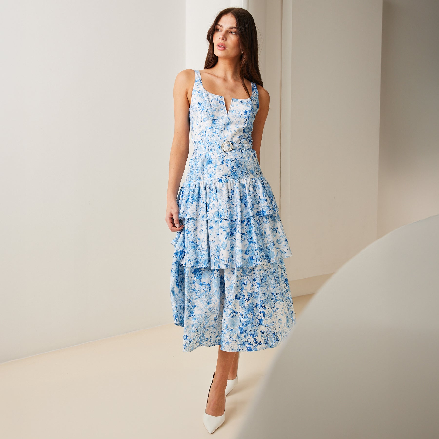 model stands wearing a blue and white floral tiered dress with crystal buckle belt and white heels.