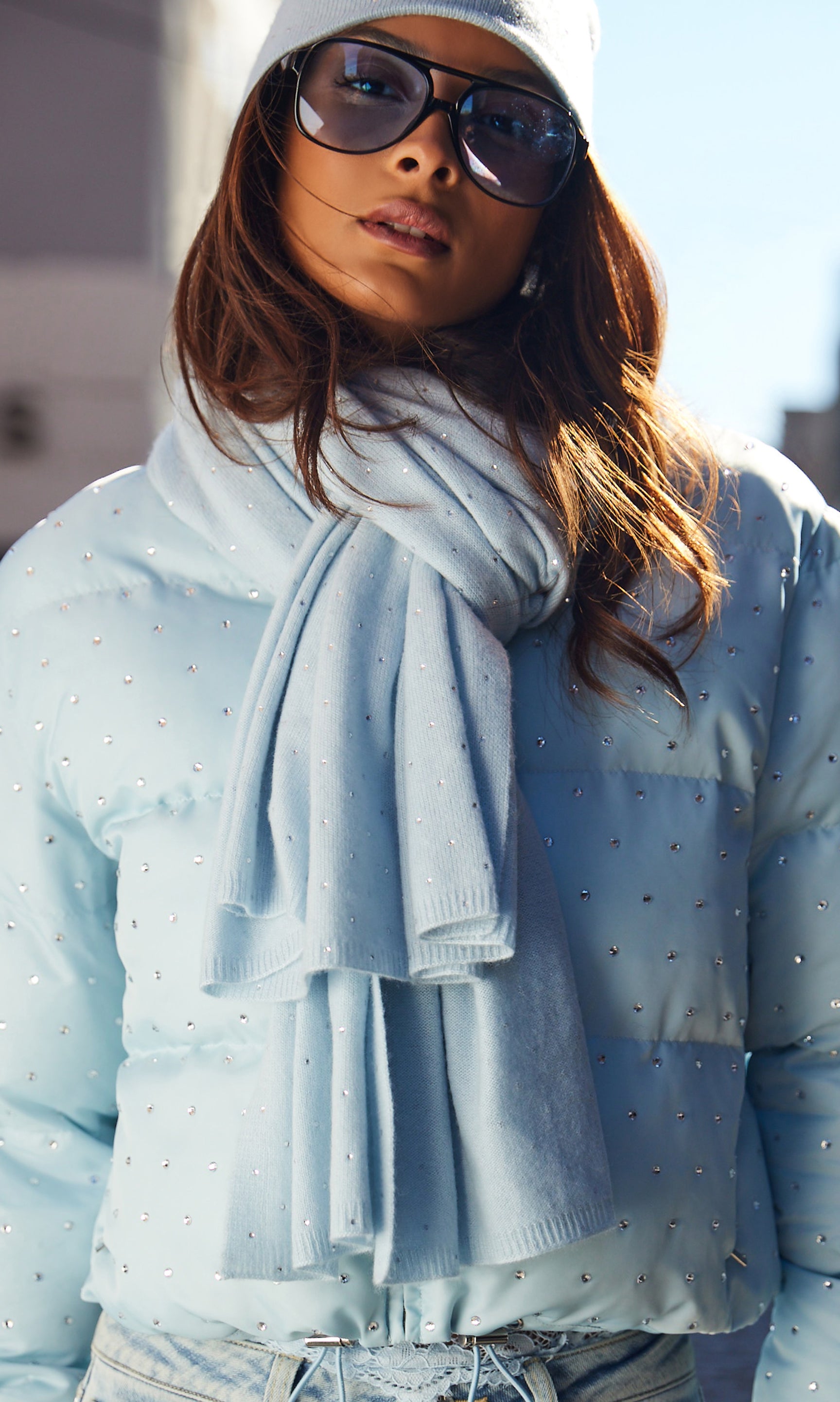 Person wearing a light blue jacket and scarf with sunglasses and a beanie.