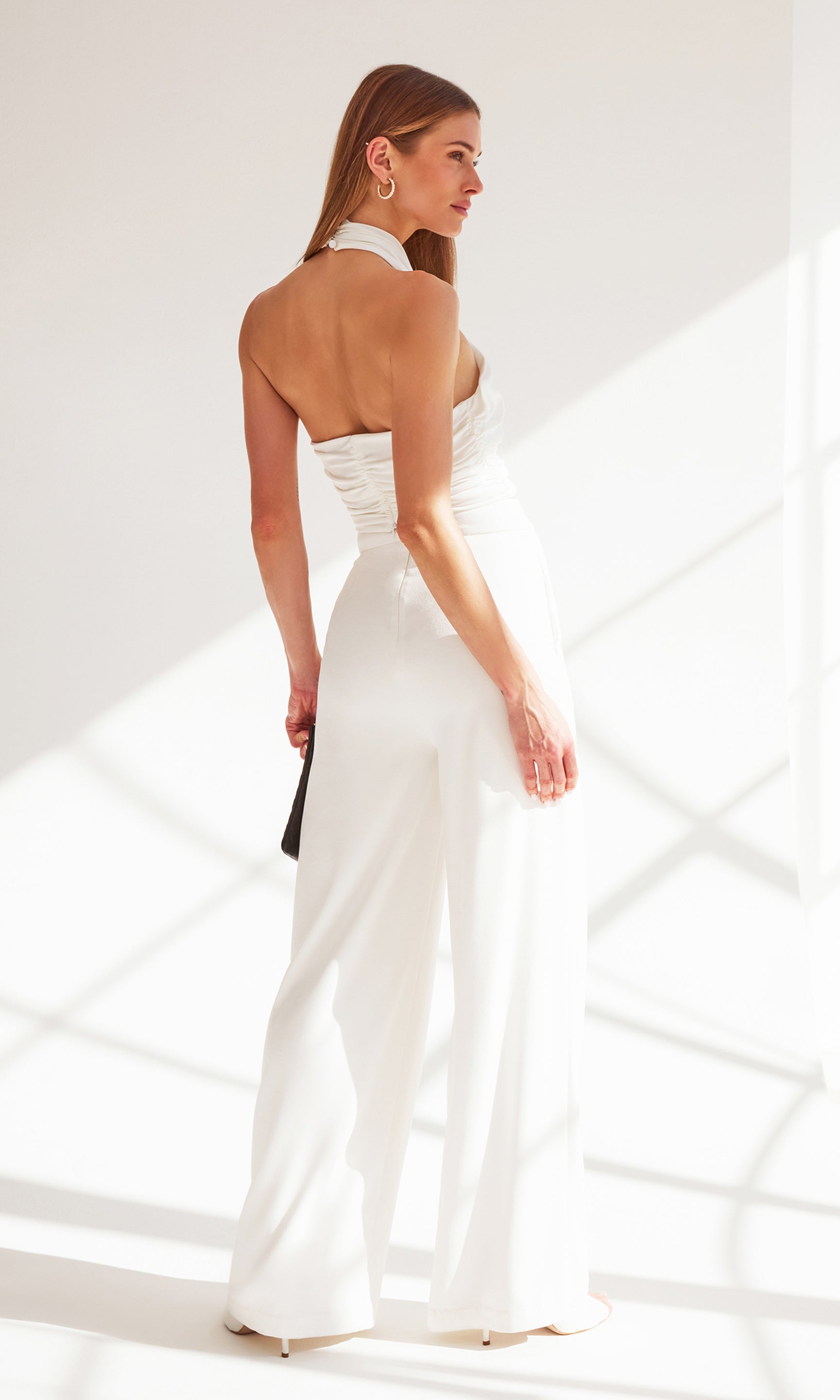 back view image of model standing near a window wearing the Edith halter top in white with white trousers, holding a black clutch purse
