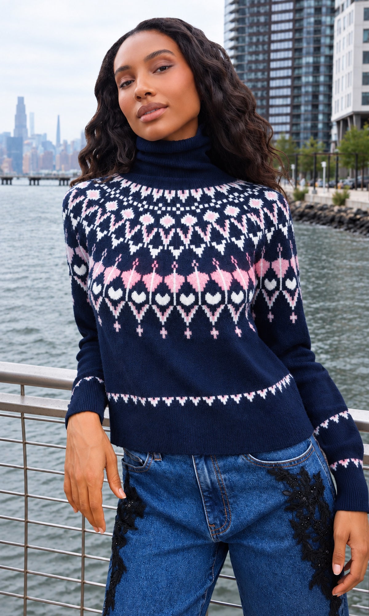 A woman wearing a navy blue turtleneck sweater with a pink and white geometric pattern, paired with blue jeans featuring black lace details.