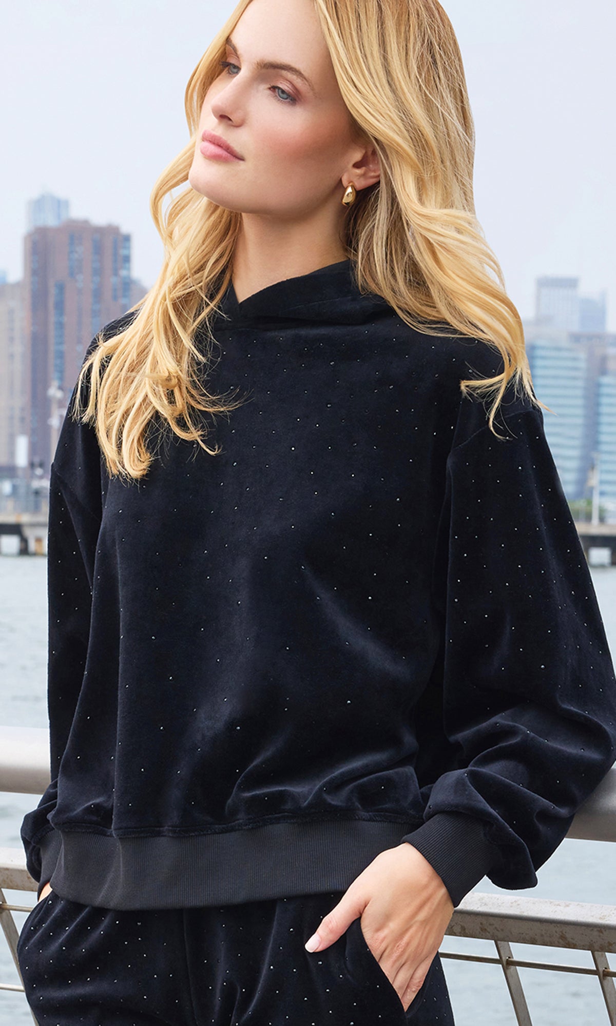 A woman wearing a black velvet long-sleeve sweatshirt with subtle silver glitter speckles, posed outdoors with a city skyline and water in the background.