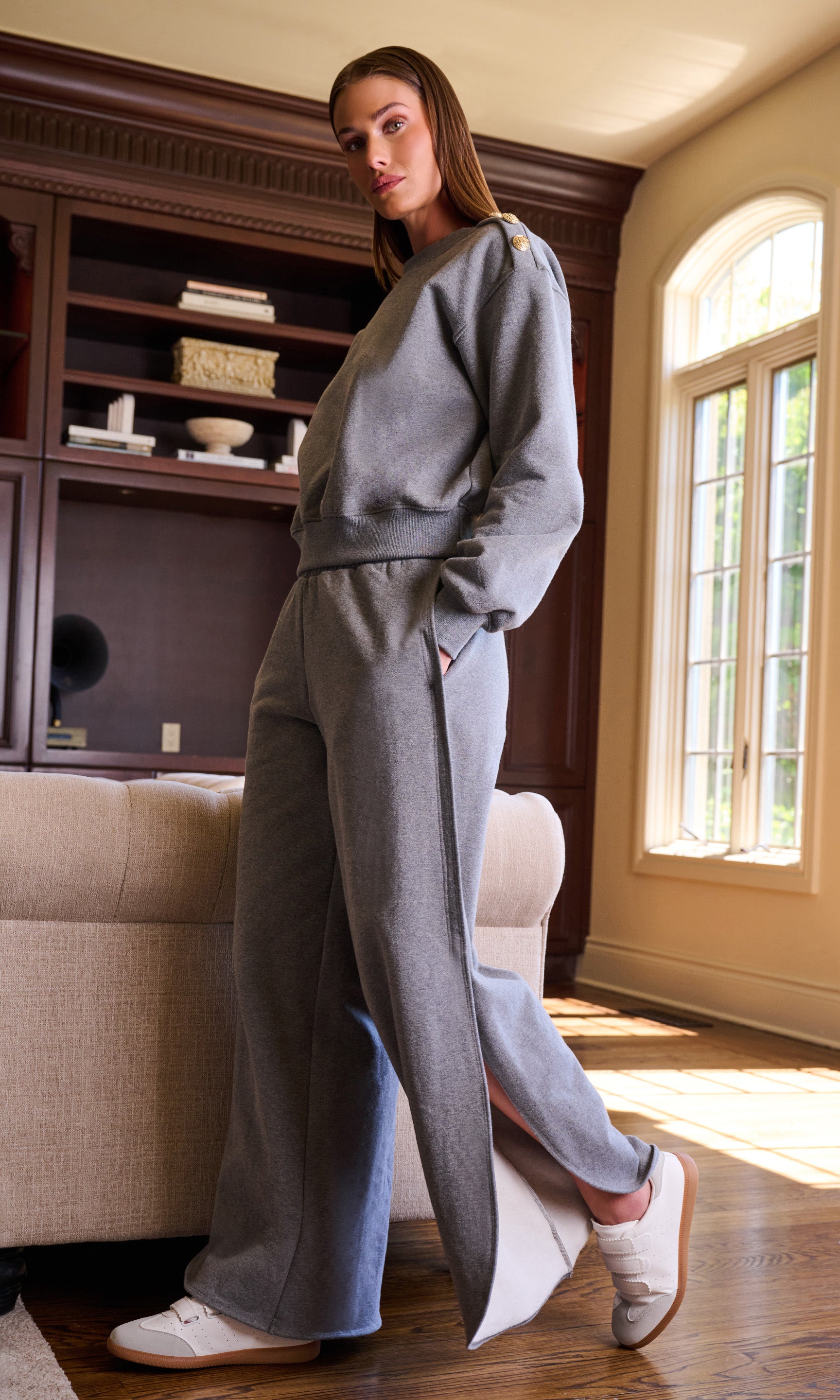 Model wearing a matching gray lounge set consisting of a crewneck sweatshirt and wide-leg pants with side slits, in a warm, sunlit room with a wooden bookcase and window.
