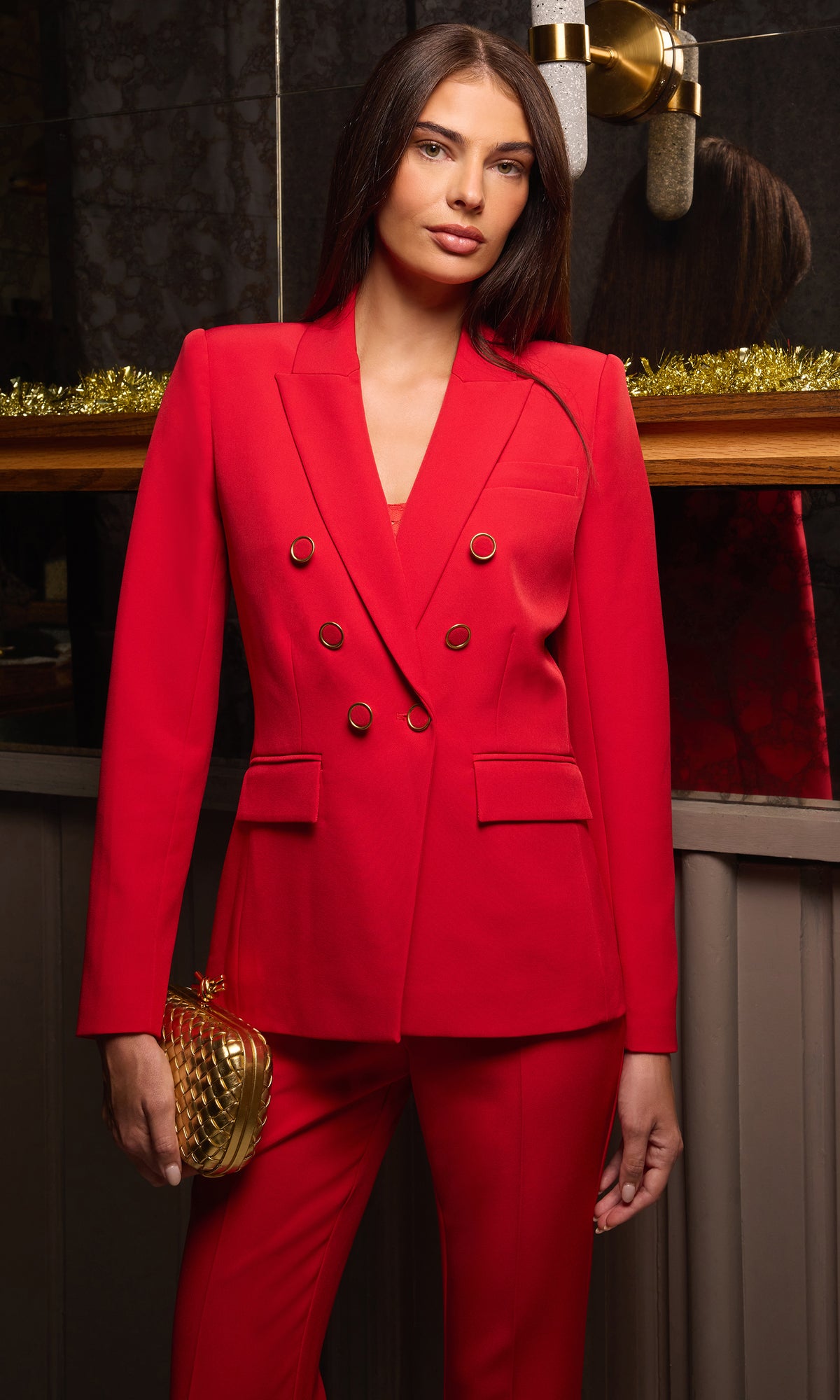 A woman modeling a bright red double-breasted blazer and matching trousers, holding a textured gold clutch in a dimly lit indoor setting.