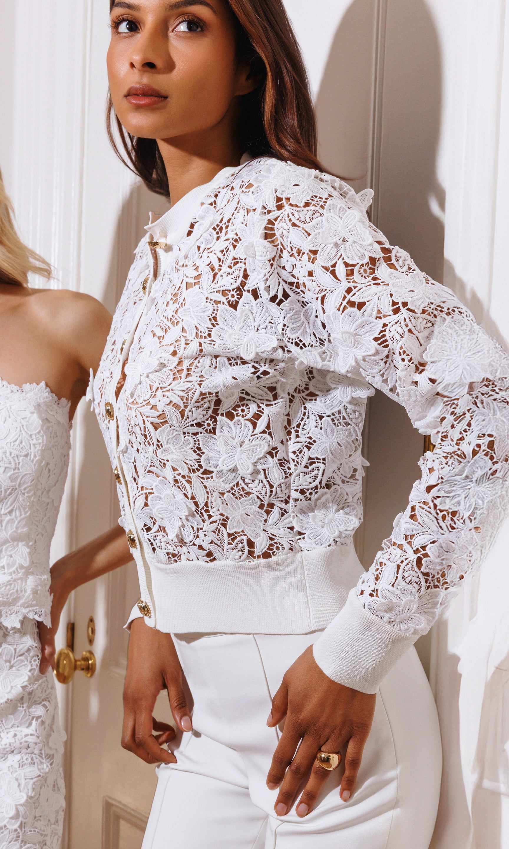 Woman wearing a white lace cardigan and pants in a well-lit room.