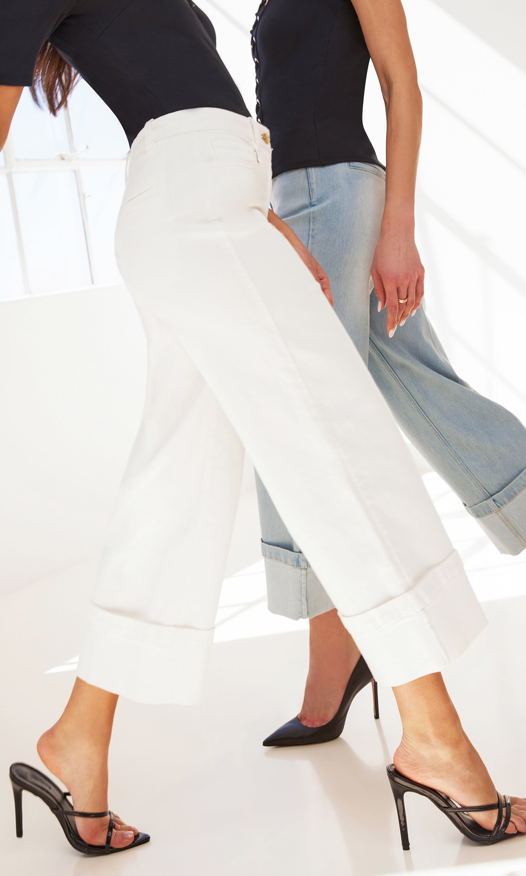 image of two models posedly walking by each other, both wearing the Eros jeans in different colors, both are paired with black shirts and black heels
