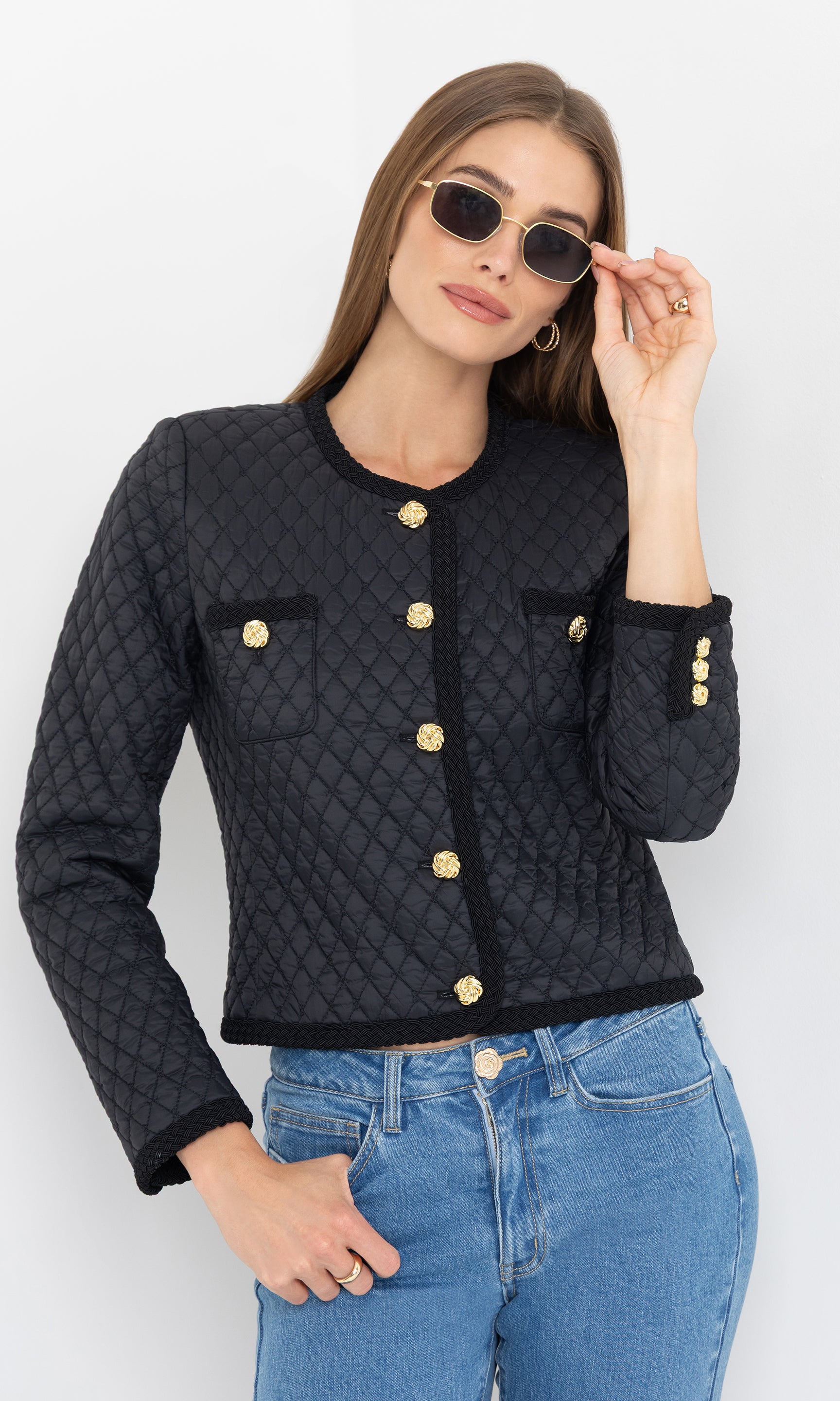 Wearing sunglasses, model poses confidently in the black Everie Quilted Jacket with gold button details and blue jeans.