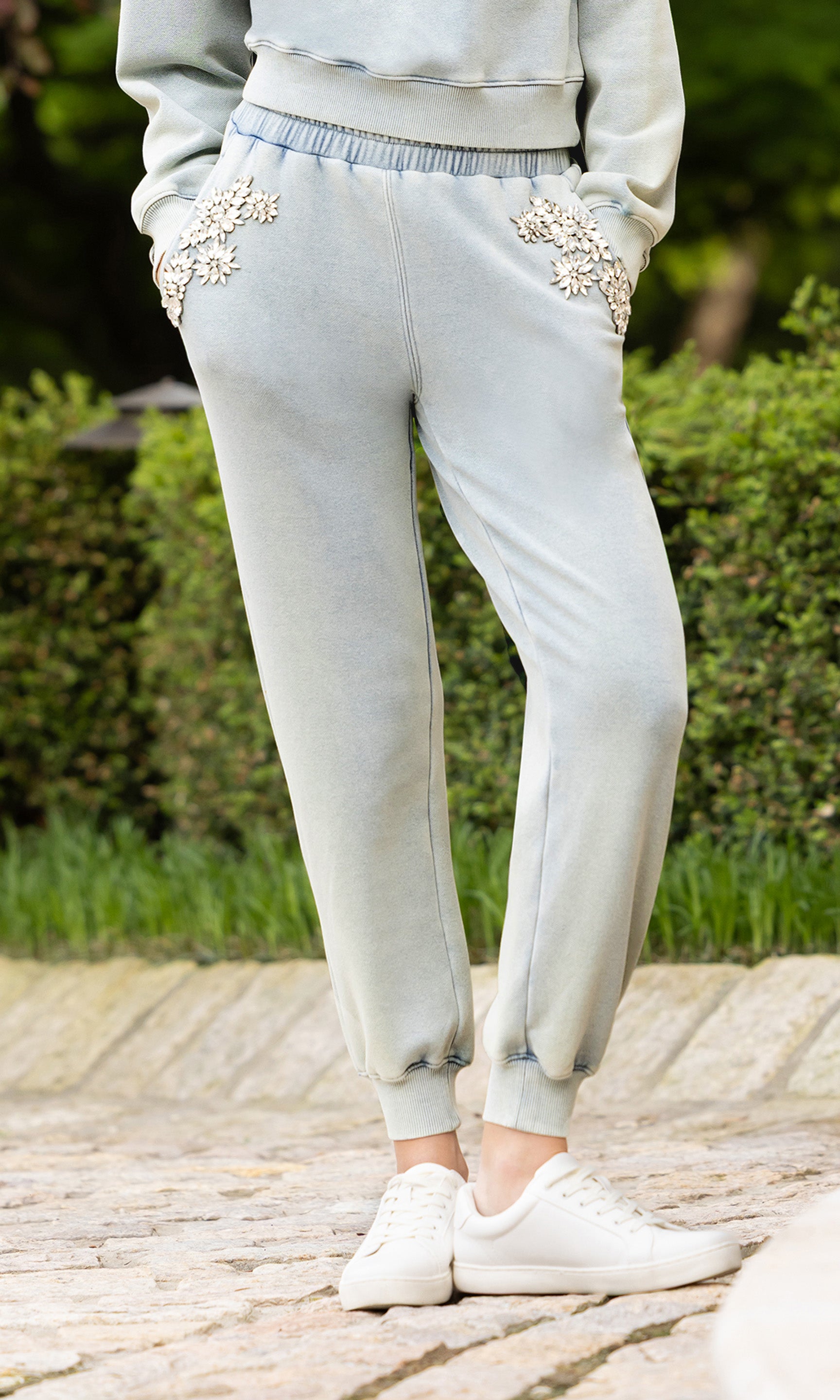 cropped in image of model in a garden patio showcasing the Suki sweatpants in a light blue washed denim color with crystal embellishments on the front pockets paired with a matching crew neck and white tennis shoes