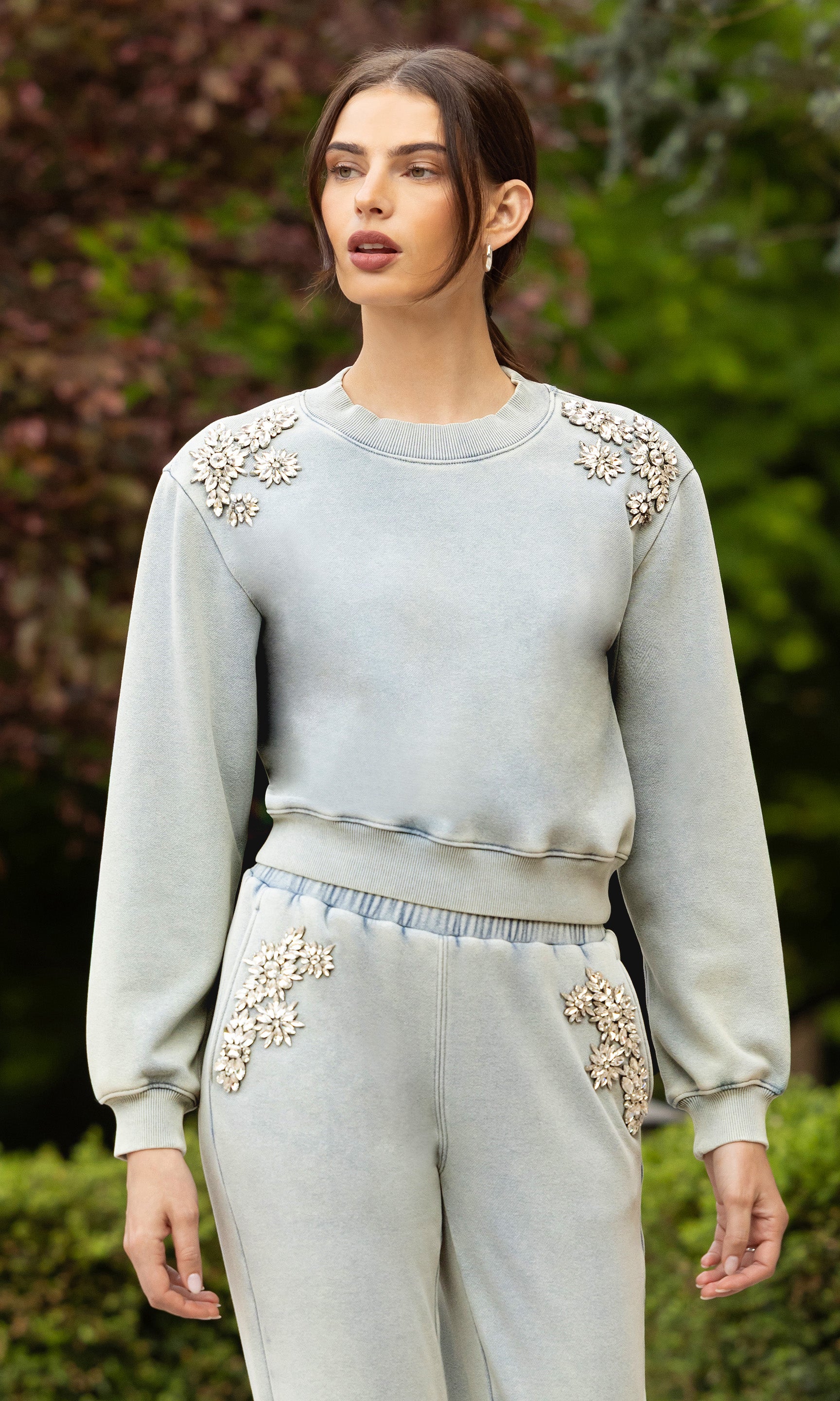 image of model standing outdoors posed, wearing the Julie sweatshirt in a light blue washed denim color with crystal embellishments on the shoulder, paired with matching sweatpants