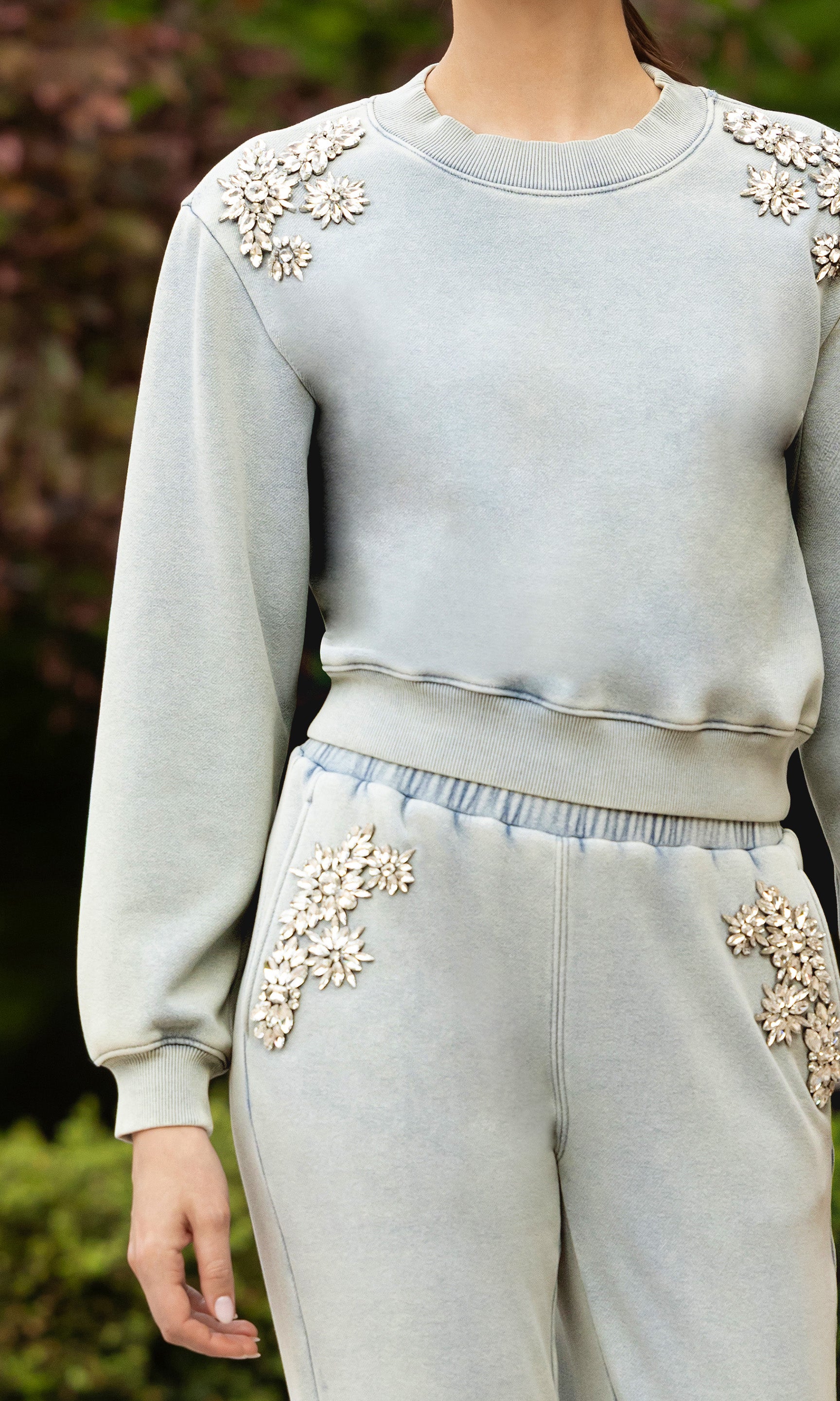 close up of image of model standing outdoors posed, wearing the Julie sweatshirt in a light blue washed denim color with crystal embellishments on the shoulder, paired with matching sweatpants
